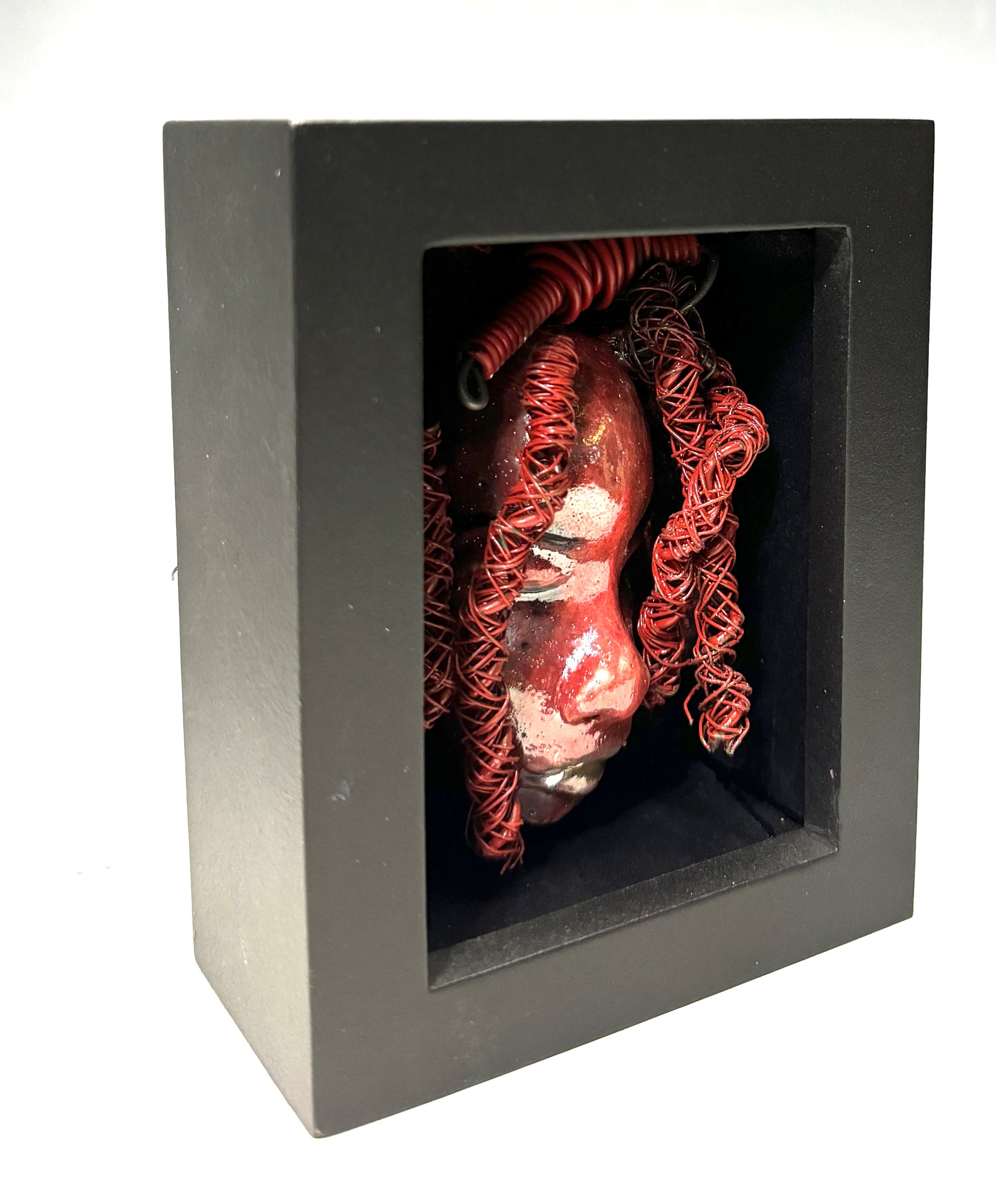 Discover your daring side with Rose - a striking, one-of-a-kind mixed media mask that has been raku fired and placed in a 4" x 4" black shadowbox. Featuring over 25 feet of 16 and 24 gauge wire as hair, this bold and unique piece will add a touch of adventure to any space. Embrace the unconventional and stand out from the ordinary with Rose, the statement piece that will elevate your decor with its fiery presence.