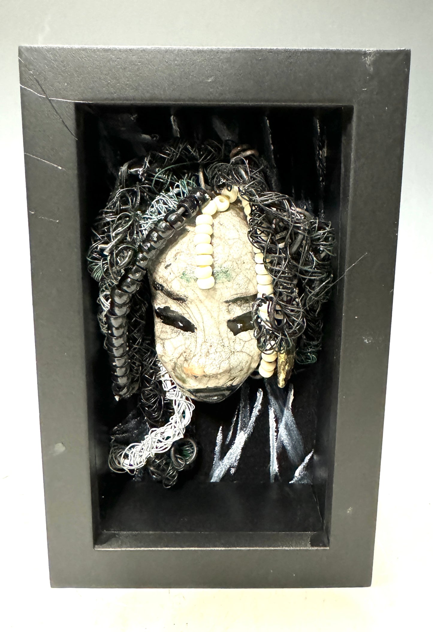 Experience the thrill of Nache: A captivating mixed media mask crafted with the daring raku firing technique. Masterfully designed with a striking dual-toned white crackle finish and skillfully woven hair using 16 and 24 gauge wire. Adorned on a hand-painted canvas against a breathtaking landscape backdrop, this piece is elegantly displayed in a sleek 5" x 7" black shadowbox, shielded by a glass cover.