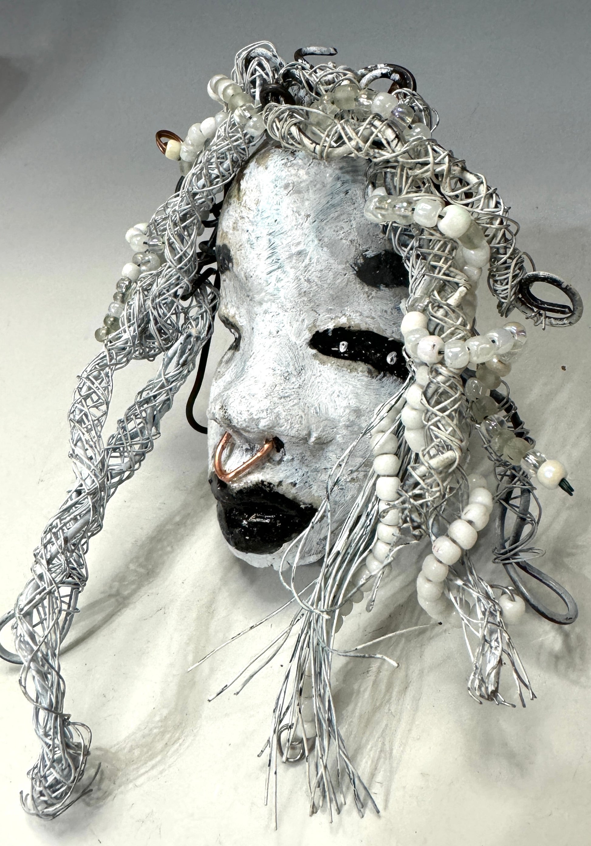 Juanita is a classic 4" x 4" 4.2 oz. Raku-fired mask designed with a white-hued complexion. Her hair is a long 15+ feet of 16-gauge wire, adorned with delicate beads. The portrait of her is set outdoors amongst a tree, making her an ideal present for art and nature admirers, as well as a perfect addition to any outdoor décor.