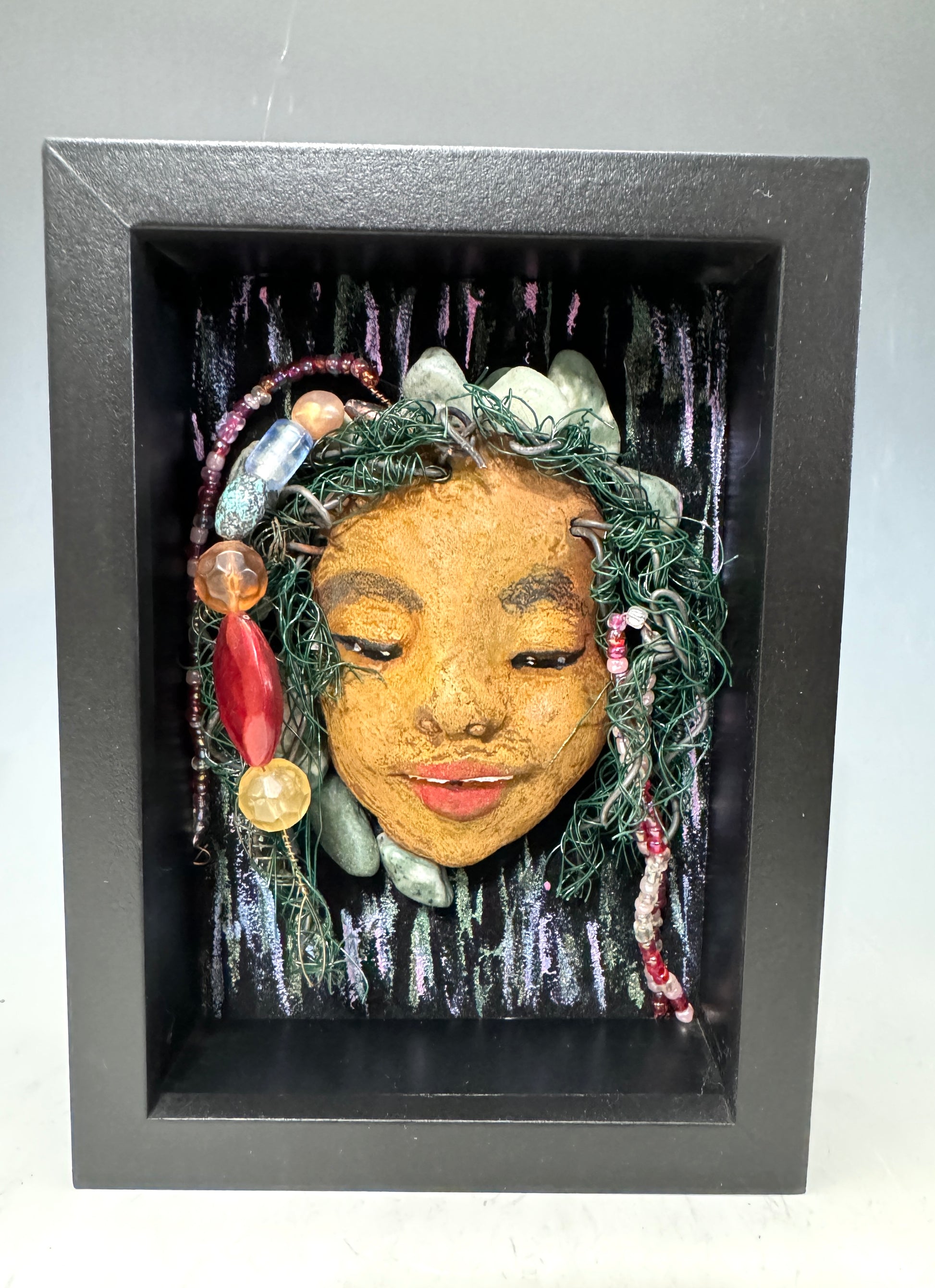 Presenting Haley: A captivating mixed media mask made with a raku firing technique. Meticulously crafted features include a unique two-tone honey brown complexion, subtle Ruby red lips, and hair intricately woven with 16 and 24 gauge wire. This piece is mounted on a hand-painted canvas, featuring a beautifully landscaped background, and elegantly displayed in a glass covered 5" x 7" black shadowbox.