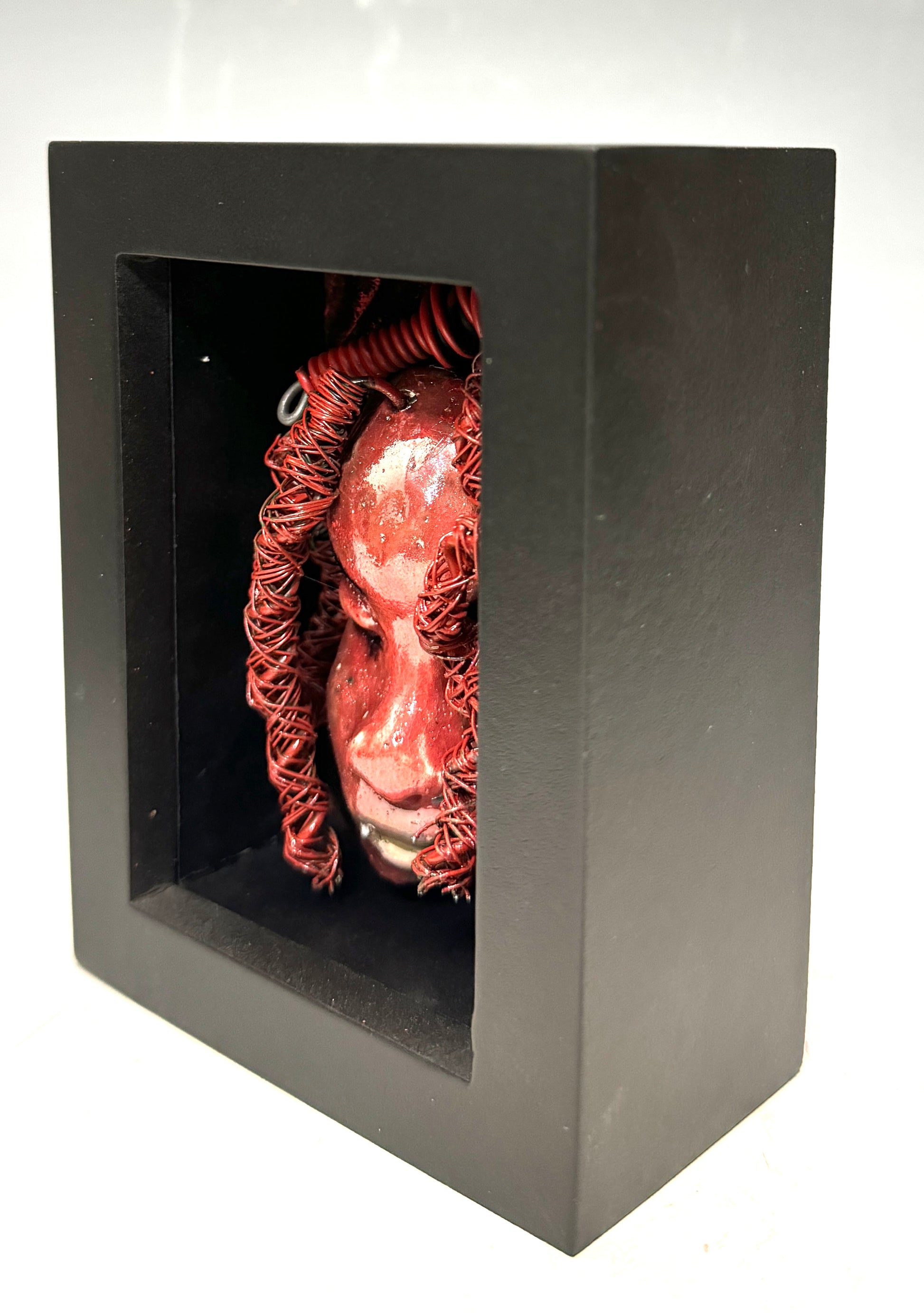 Discover your daring side with Rose - a striking, one-of-a-kind mixed media mask that has been raku fired and placed in a 4" x 4" black shadowbox. Featuring over 25 feet of 16 and 24 gauge wire as hair, this bold and unique piece will add a touch of adventure to any space. Embrace the unconventional and stand out from the ordinary with Rose, the statement piece that will elevate your decor with its fiery presence.