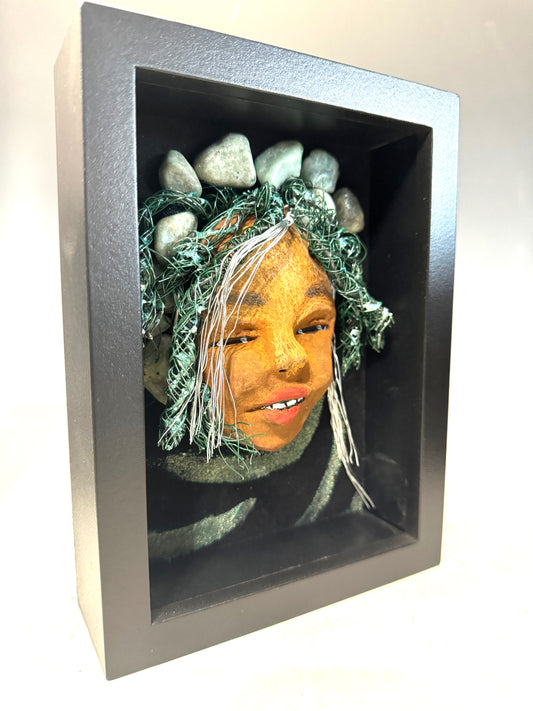 Introducing Brandie, a mixed media sculptured raku fired mask. Her features include a two-tone honey brown complexion, Ruby red lips, and hair intricately intertwined with 16 and 24 gauge wire. She is mounted on a hand-painted canvas, accented by an earthy landscape background, and enclosed in a 5“ x 7“ black shadowbox.