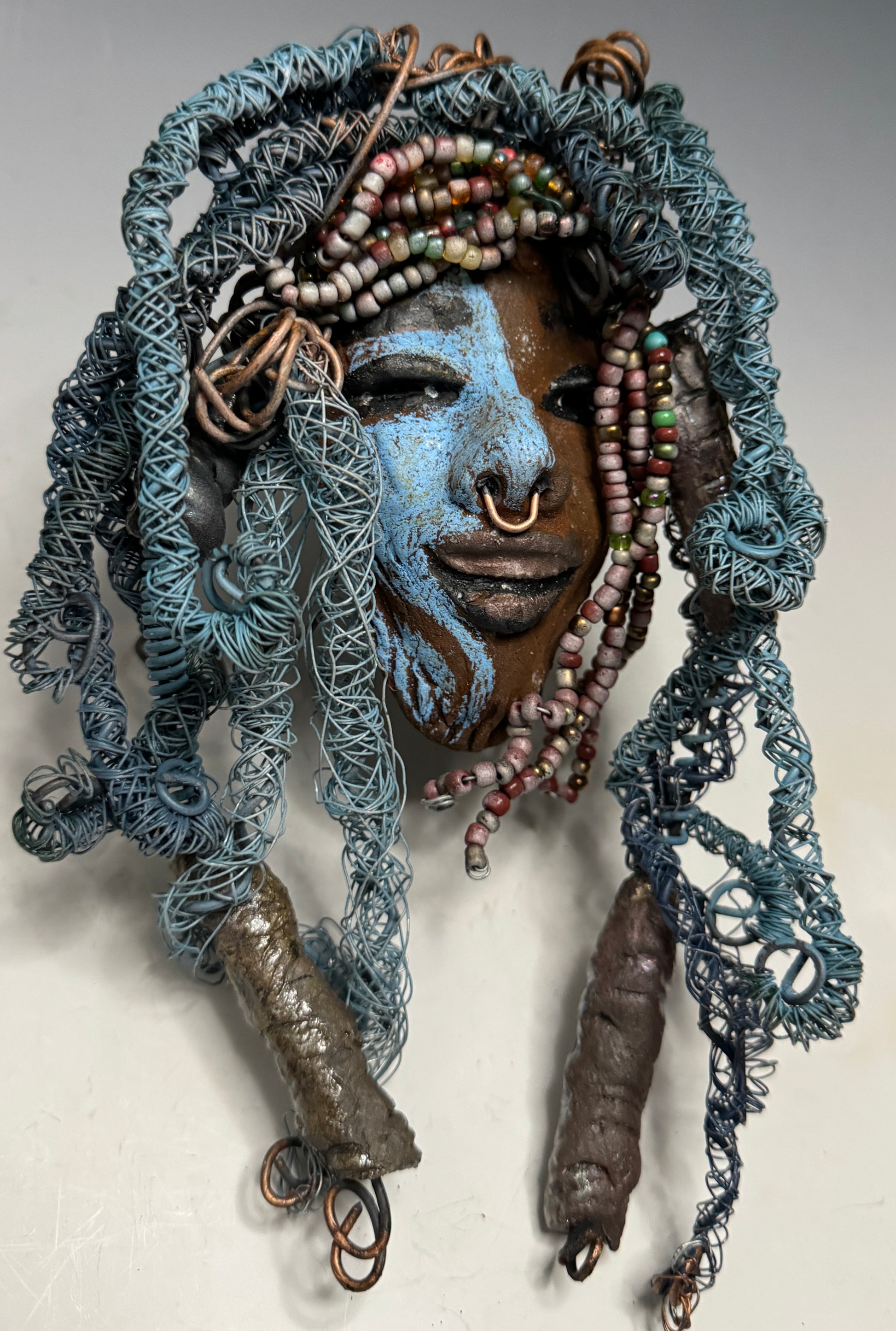 Zola is a mesmerizing raku mask with a captivating tribal face. She measures approximately 5 x 9" and weighs 7 oz. The mask is further decorated with blue hair and a copper nose ring.