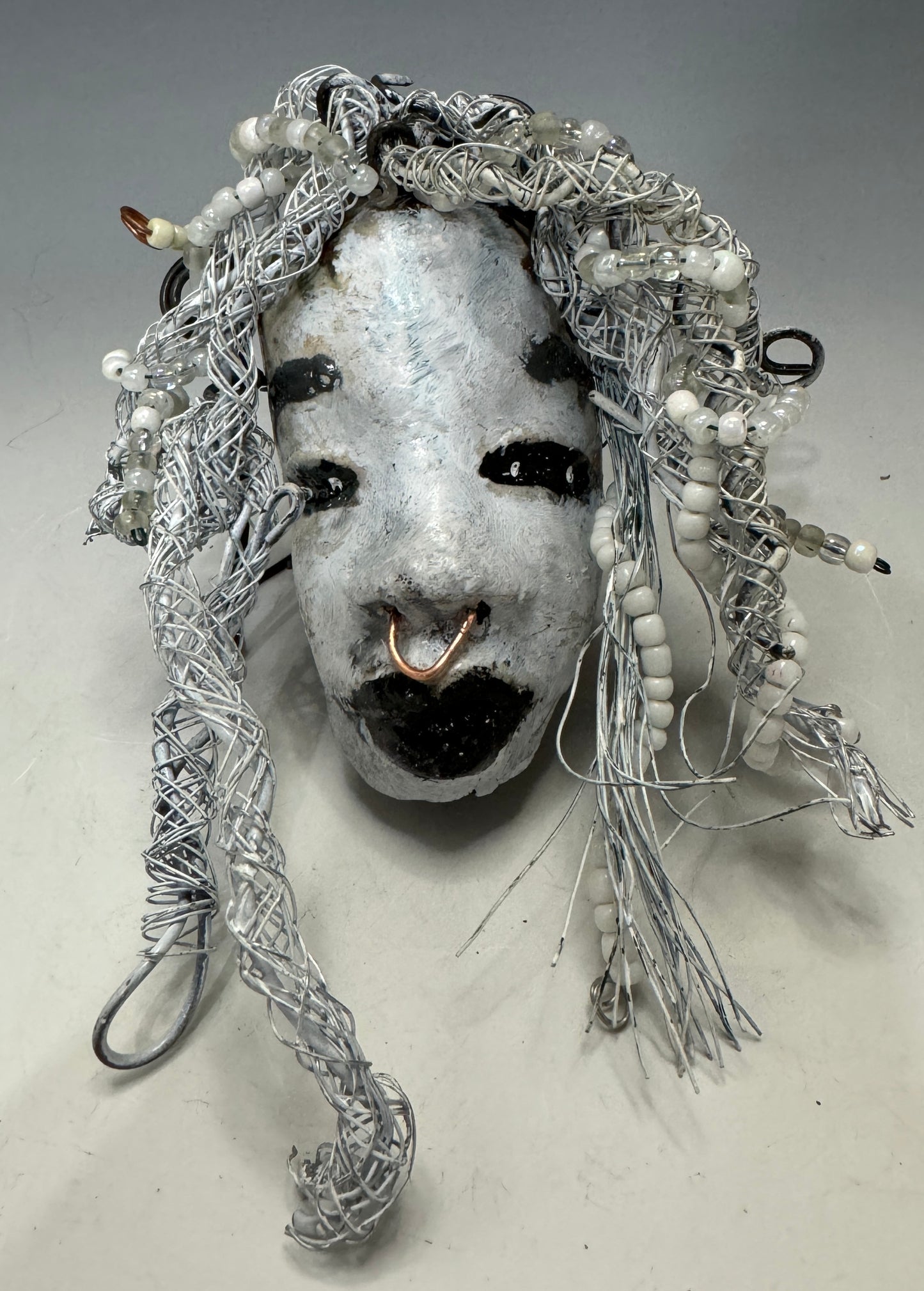 Juanita is a classic 4" x 4" 4.2 oz. Raku-fired mask designed with a white-hued complexion. Her hair is a long 15+ feet of 16-gauge wire, adorned with delicate beads. The portrait of her is set outdoors amongst a tree, making her an ideal present for art and nature admirers, as well as a perfect addition to any outdoor décor.