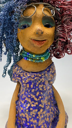 Cristy is 9" x 6" x 5.5", weighs 2.5 lbs, and has a honey brown complexion with reddish brown lips. Her long hair is adorned with red and blue color and 80 feet of 24 and 16 gauge twisted wire. She wears an antique glazed dress featuring a spotted blue and gold pattern. Cristy is completed by her eyeglasses which sit upon her forehead, inspiring hope and sparking conversations. Visit us for questions