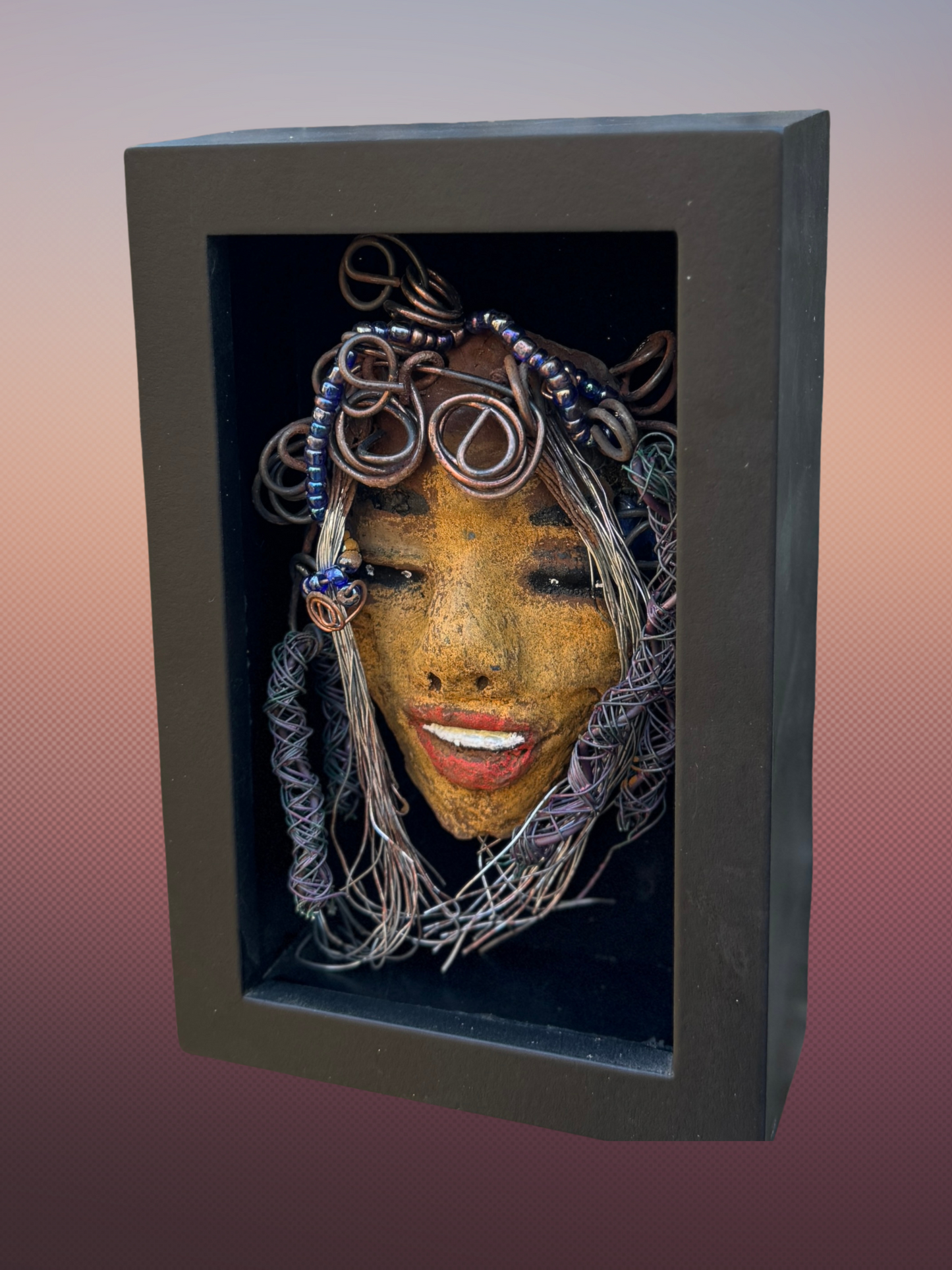 Agnes is a "4 x 6" raku mask displayed in a glass covered black shadowbox, accented with a beautiful two-tone honey brown complexion. Her intricate hair, made of 16 and 24 gauge wire, measures over 50 feet in length. With a contagious smile, Agnes adds a touch of elegance to any home decor.