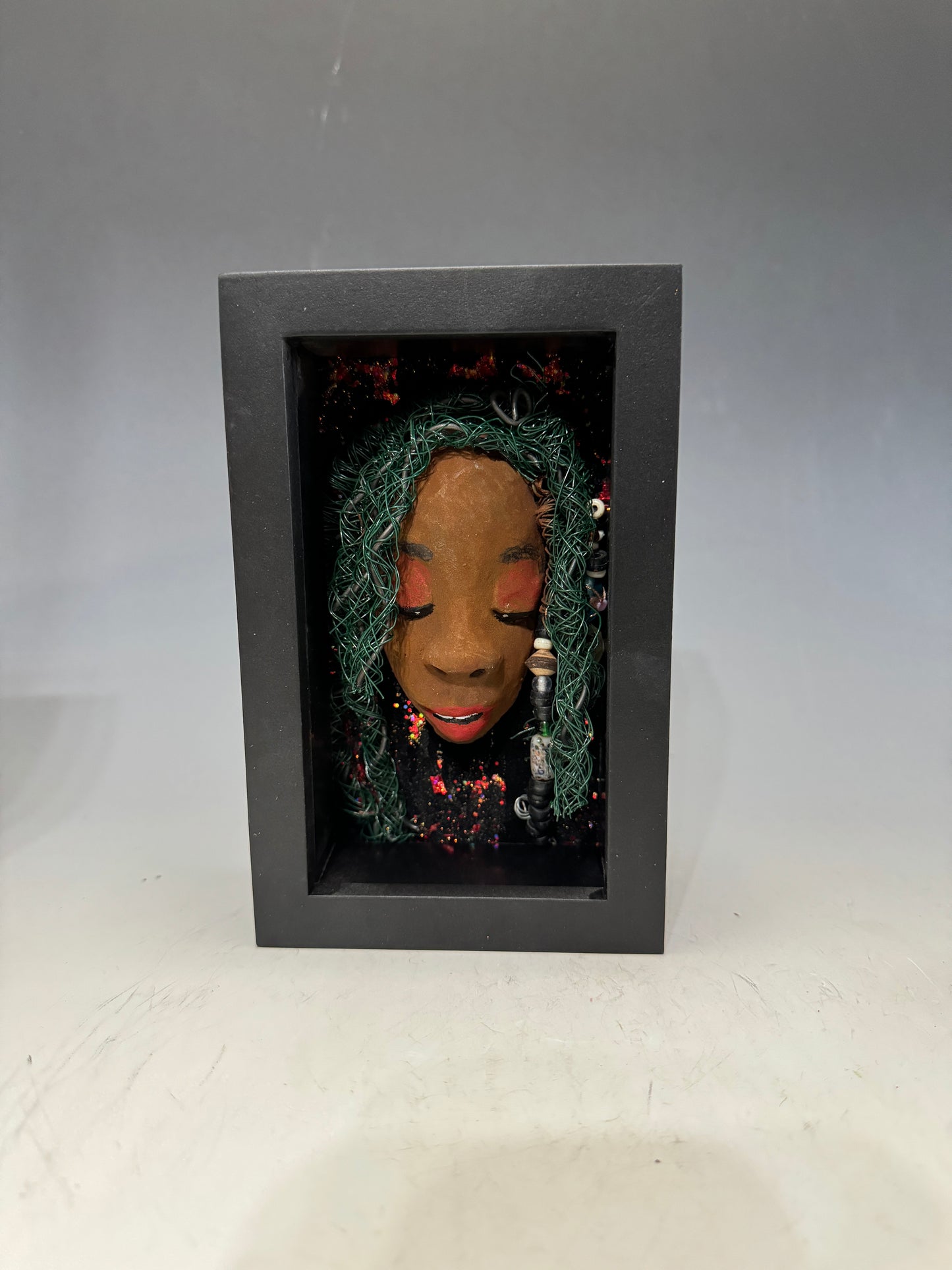 Marjorie is a mixed media mask crafted with the raku firing technique, displaying carefully detailed characteristics such as honey brown skin, Ruby red lips, and intricately woven hair made from 16 and 24-gauge wire. This stunning piece is mounted on a hand-painted canvas, surrounded by a beautifully landscaped background, and elegantly displayed in a glass-covered 4" x 6" black shadowbox.