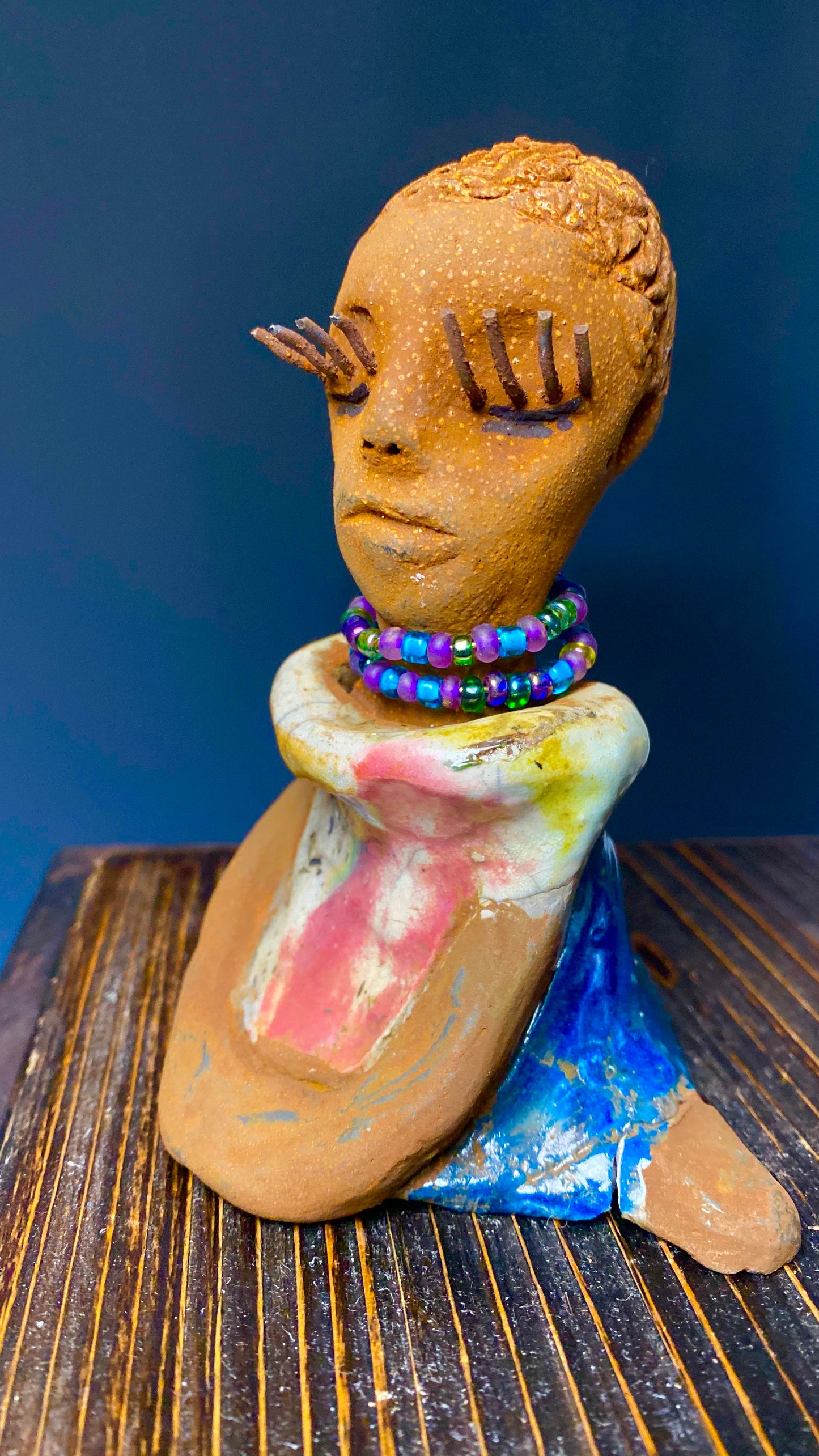 Meet Little Riley! Riley stands 5" x 5”x 3" and weighs  11 ozs.  Riley has a lovely multicolored glossy dress with a matching beaded necklace.  She has long lashes!  Riley appears to sit in a yoga pose. Her long arms rest at her side. Riley is a great starter piece from the Herdew Collection!