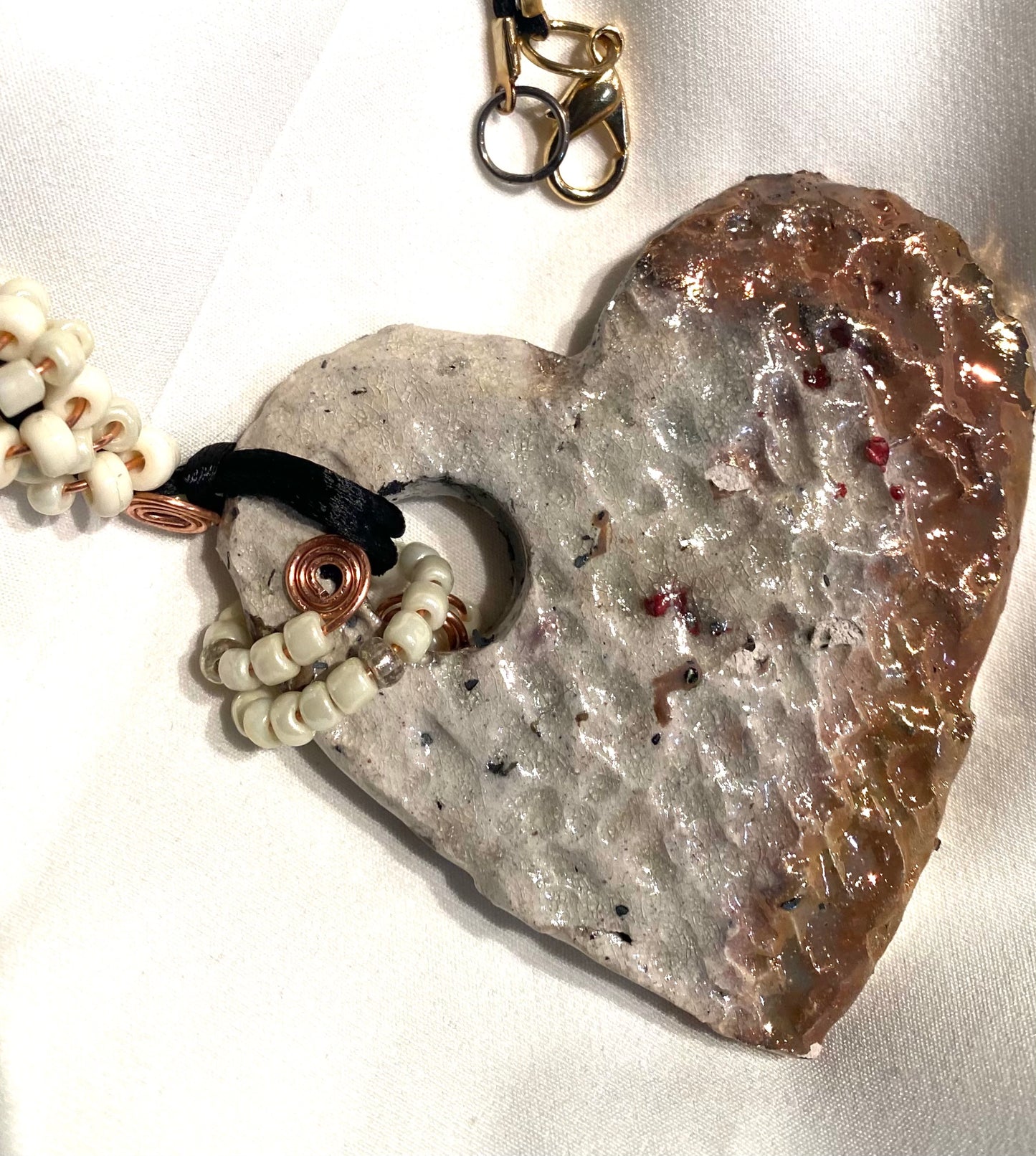 Have A Heart ! Each heart pendant is handmade with love! It is 3" x 3" and weighs approx. 3ozs. It has white metallic raku glazes that renders a unique translucent  patina. The heart is textured and holds a  spiral of white pony beads on copper wire.  This pendant has a nice 12"black rattail cord!