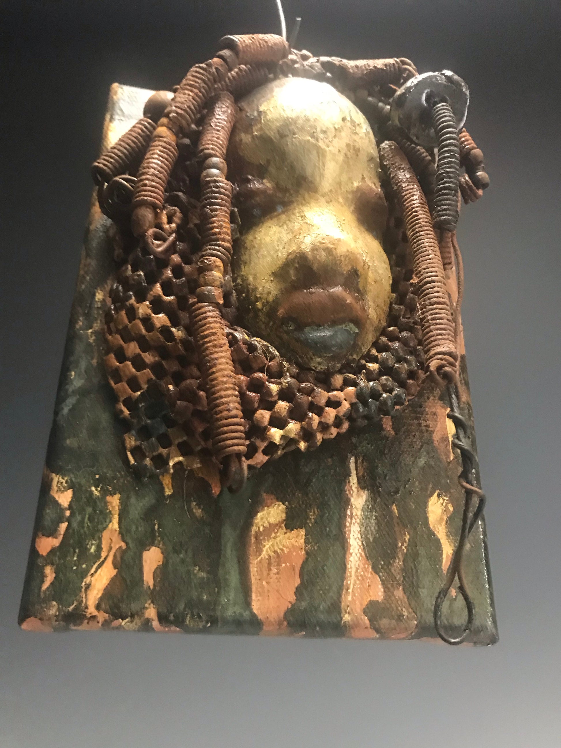 "I started making mask after seeing an authentic African Mask collection at the Smithsonian Museum of African Art. I was in total awe. Dillan was inspired by my visit there".       Dillan is mounted on a painted 6"x 4"x 2" canvas. She weighs 15 ounces.     Her face is formed with hand coiled wire, multiple raku, and wood beads, matte textured  and brown cloth.     Dillan's face beams with  almond  green glaze and rust blue brown lips.     Dillan is ready to be hung.