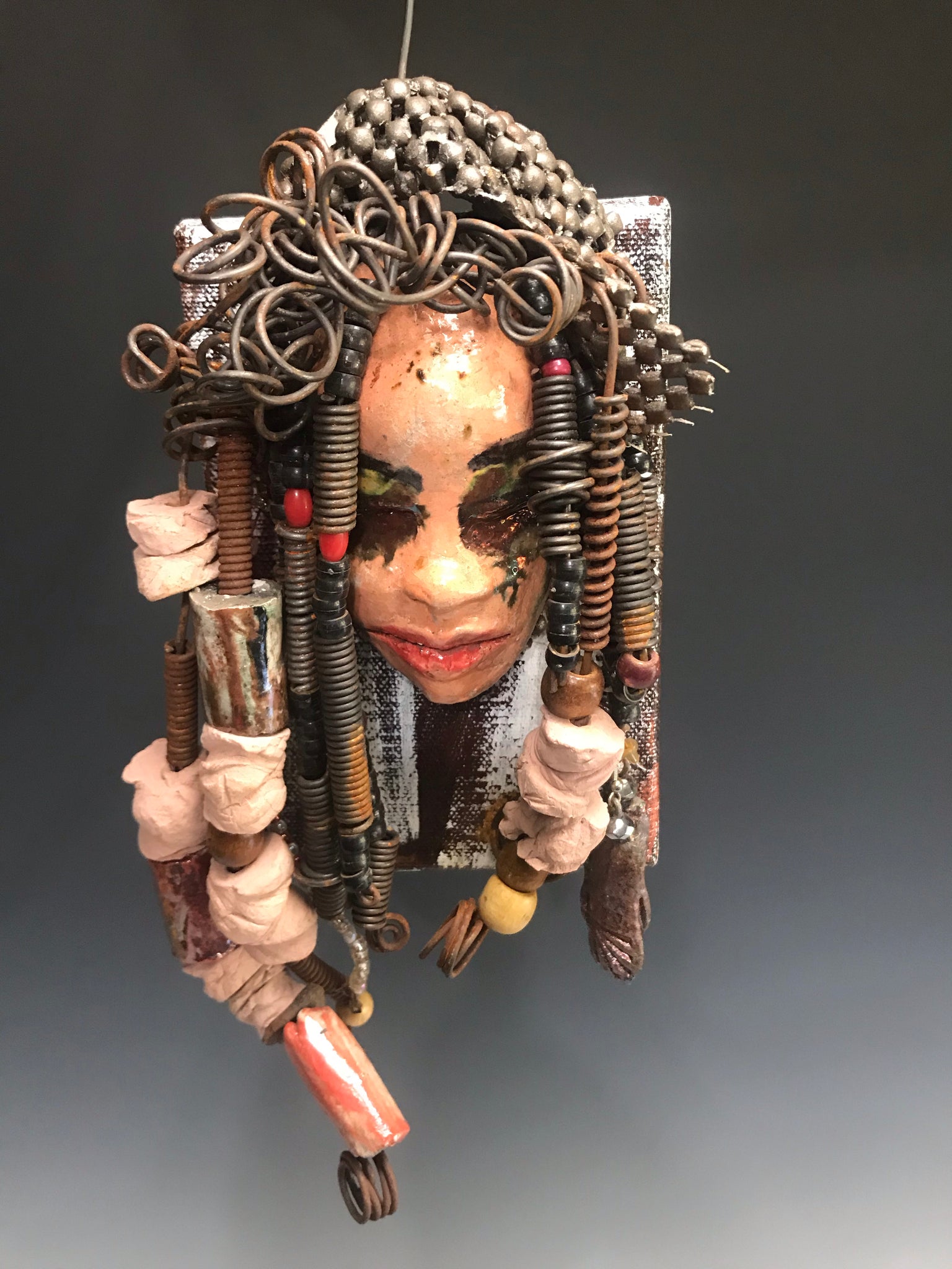      Kamara is uniquely mounted on a painted 6"x 4"x 2" canvas. It weighs 1.6lbs.     Her face is formed with hand coiled wire, raku beads, textured brown cloth, and multi color raku beads.     Kamara's face beams with pale pink crackle glaze and ruby red lips.  Kamara is ready to be hung in a special place!   