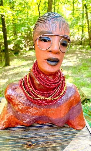 Meet Harriet! Harriet stands 13" x 11" x 5" and weighs 5 lbs. She wears a coral striped dress with a string of ruby red and gold beads. Harriet has a honey brown rust complexion  and distinct eyes. Her eyes  are slightly opened underneath her glasses... She has mixed black and copper streaks of straight corn rolls of clay hair. She has a straight look of concern as to what is about to take place.