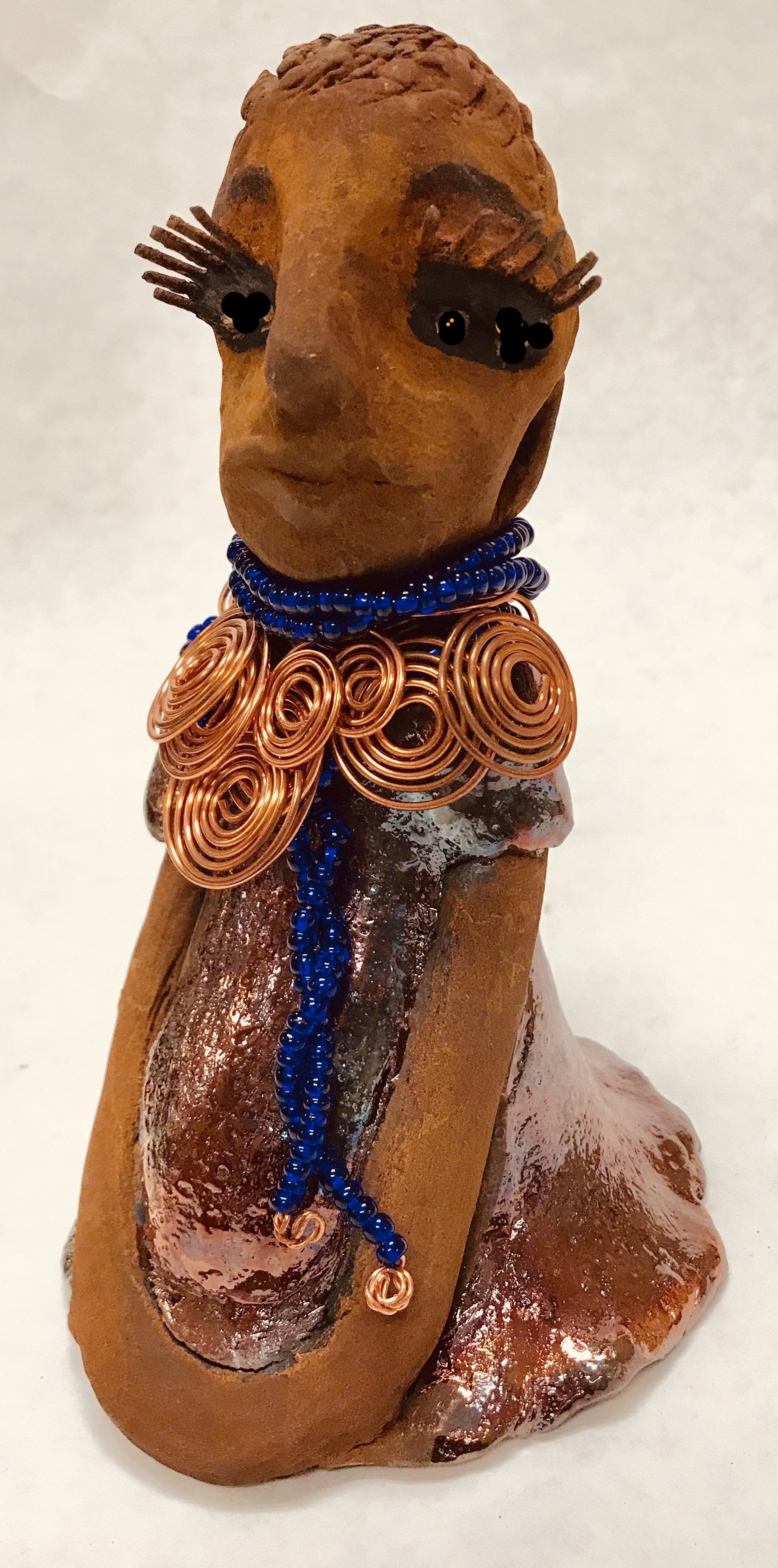 Tattayana stands 8" x 4.5" x 5" and weighs 1.02  lbs. She has a lovely honey brown complexion with reddish brTattayanaown lips. She has a short braided hairstyle.  Tattayana has a colorful metallic antique copper glazed dress. She wears spiral copper wire necklaces on top of an aqua blue beaded collar.  Tattayana has her long loving arms rest at her side. With eyes wide opened,Tattayana  has hopes of finding a new home.
