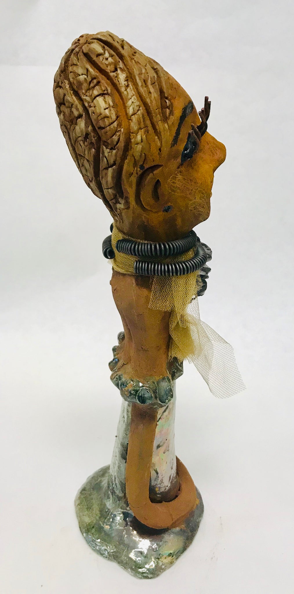 Trinity stands 14" x 4" x 4" and weighs 2.7 lbs. She has a lovely honey brown complexion with  reddish brown lips. She has a braided hairstyle.  Trinity has a colorful metallic green antique copper glazed dress. She wears a spiral wire necklaces with raku beads underneath an antique gold tulle.. With long lashes and eyes wide opened, Trinity has hopes of finding a new home.  