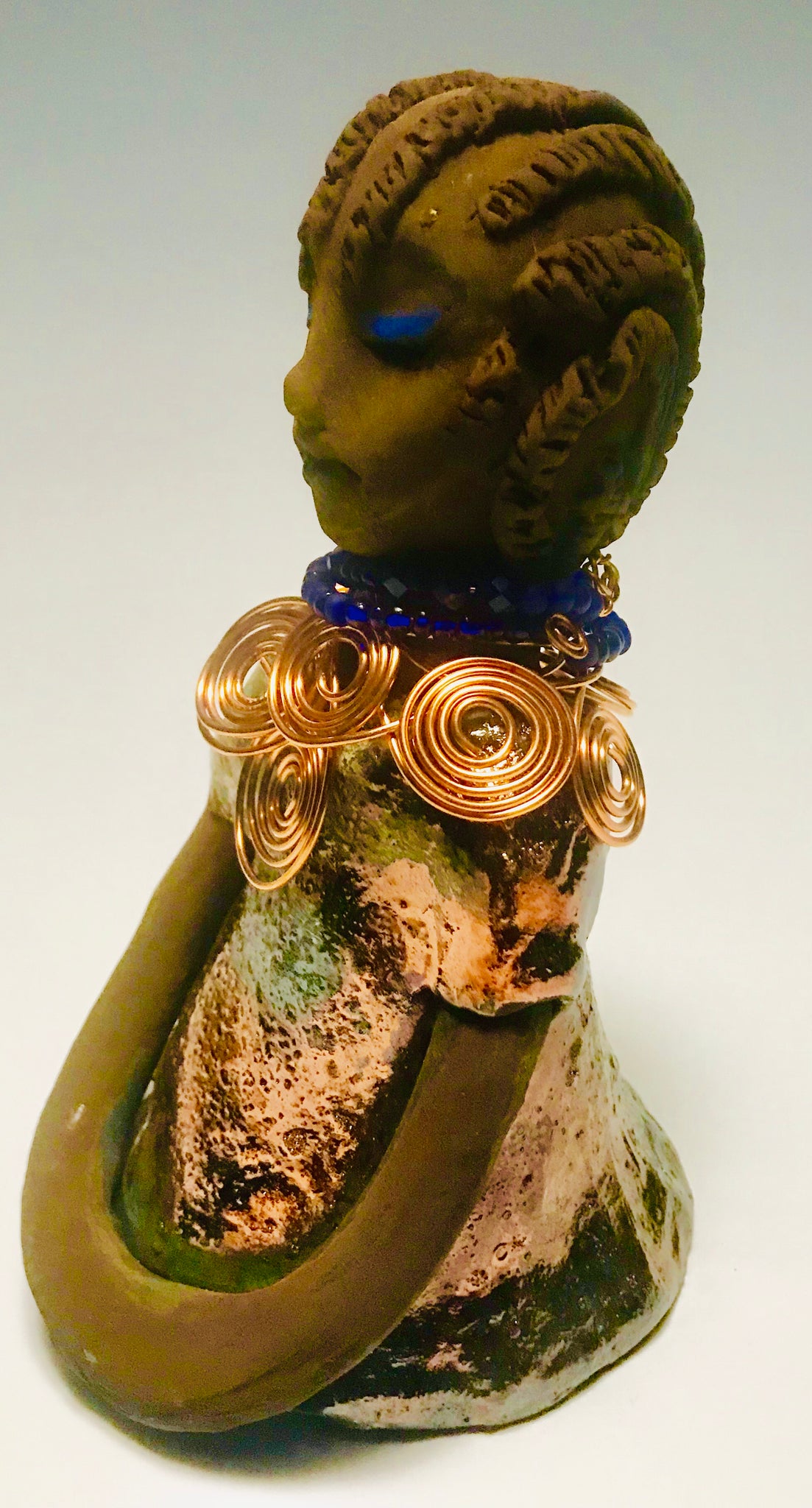 Anja stands 7" x 4" x 4" and weighs 1.01 lbs. She has a lovely honey brown complexion with  reddish brown lips. She has a braided hairstyle.  Anja has a colorful metallic green antique copper glazed dress. She wears a spiral copper wire necklace on top of a blue beaded collar. With eyes slightly opened, Anja has hopes of finding a new home.