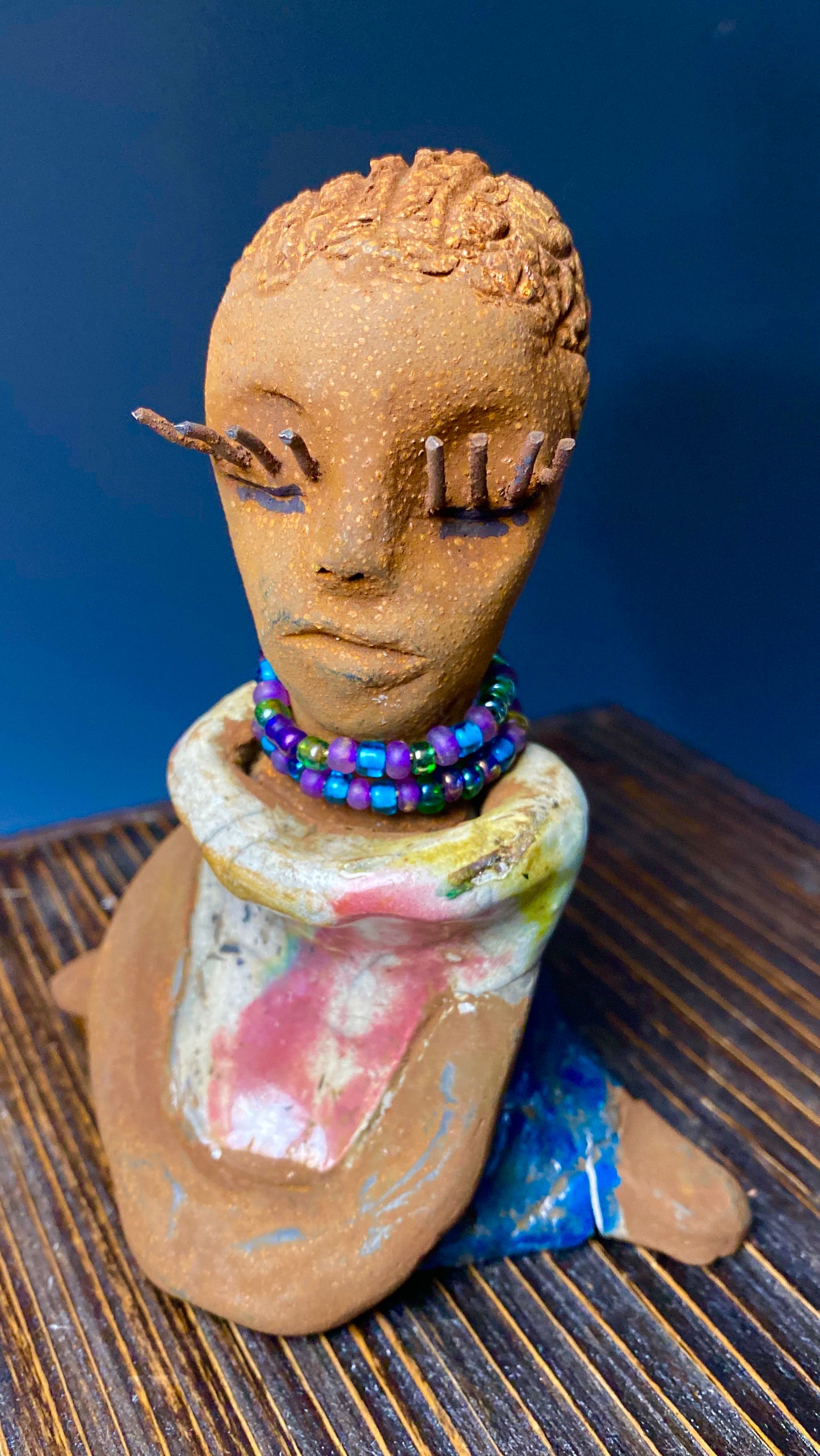 Meet Little Riley! Riley stands 5" x 5”x 3" and weighs  11 ozs.  Riley has a lovely multicolored glossy dress with a matching beaded necklace.  She has long lashes!  Riley appears to sit in a yoga pose. Her long arms rest at her side. Riley is a great starter piece from the Herdew Collection!