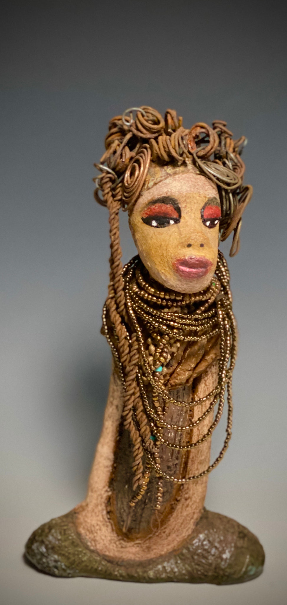 You will love having Cardi around! Cardi stands 11" x 5.5" x 3" and weighs 1.14 lbs. She has an awesome honey beige complexion. Cardi wears a dark metallic copper dress with a chocolate ruffle scarf.  She wears a multitude of amber colored hand strung beads. I do believe Cardi would like it if you gave her a space in your home!