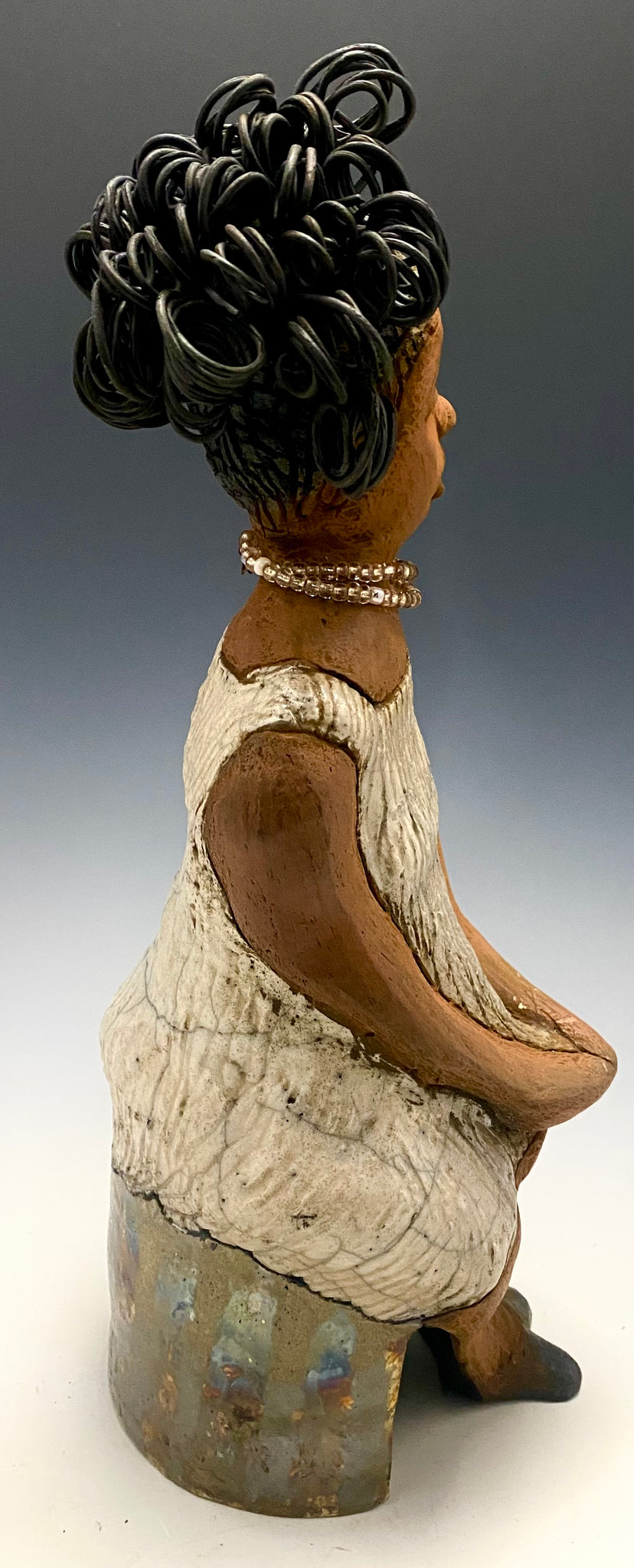 Meet Gwen!  You know someone like Gwen. One who reaps elegance and sophistication!   Gwen stands 13 " x 6" x 6" and weighs 3.07 lbs.   She has a lovely brown complexion with over 5 feet of 16 gauge spiral coiled wire hair.  Gwen's metallic off white dress is accented by a multicolored white and amber beaded necklace. Gwen’s straight face gives her a distinguished no nonsense look. Gwen sits with her hands rested as she awaits a special place to grace your home.