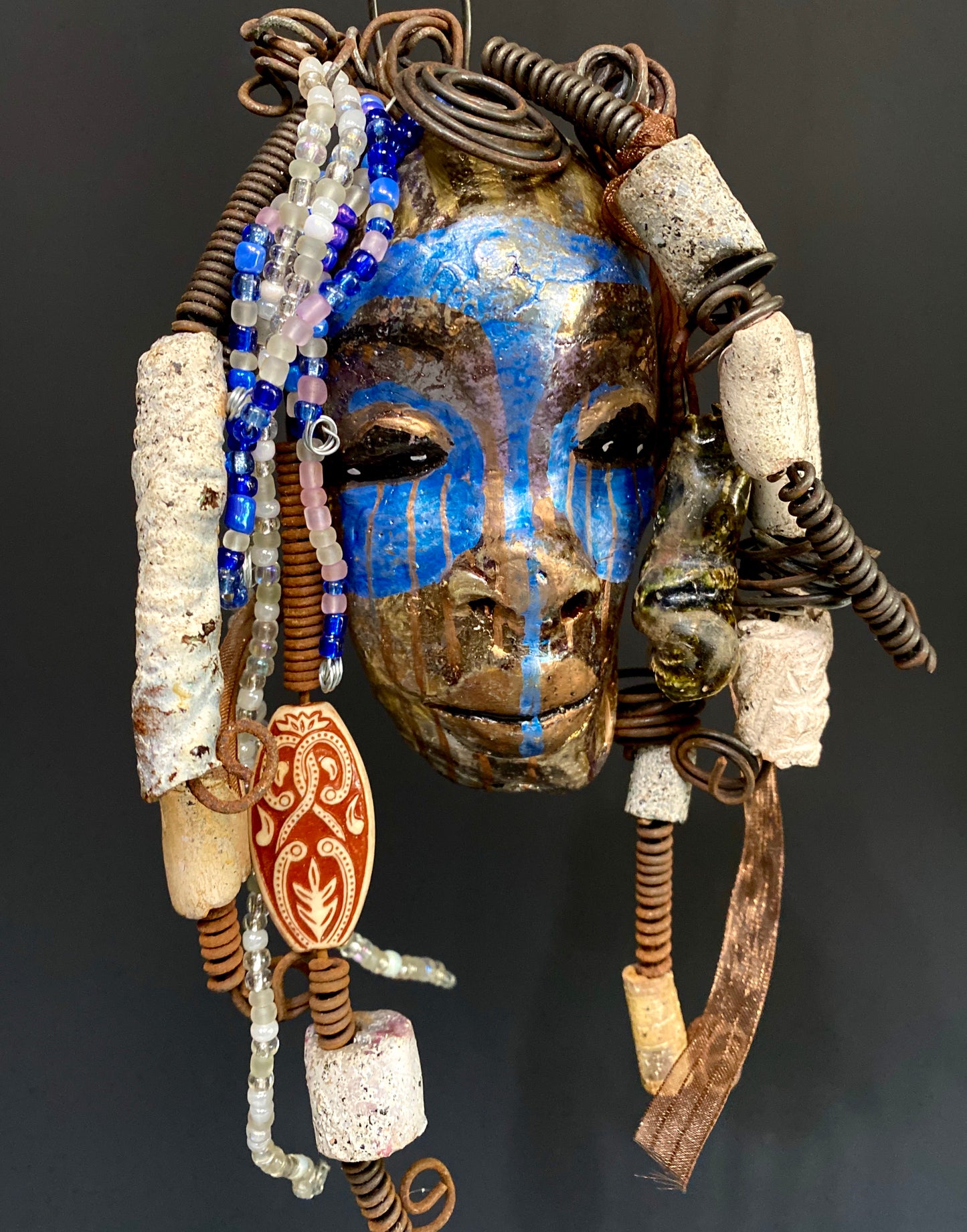 I started making mask soon after seeing authentic African artwork at the Smithsonian Museum of African Art. I was in total awe. I was inspired by my visit there..  Haze has a multi colored blue tone  metallic copper complexion. She is 7" x 5" and weighs 10 ozs. Haze  has  over 50 mini beads and handmade raku beads. Haze has over 4 feet of 16 gauge wire hair.