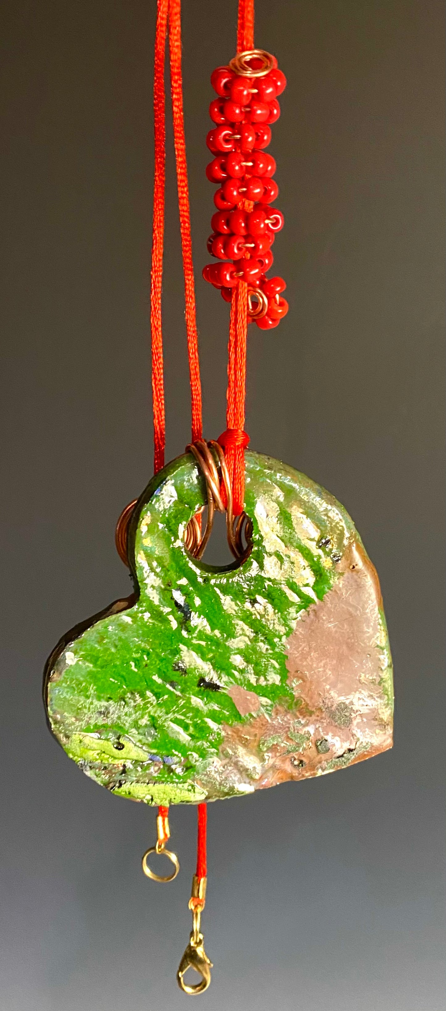 Have A Heart! Each heart pendant is handmade with love! It is 3"x 3"and weighs approx. 3ozs. This pendant has a emerald green and gold metallic raku glazes that renders a unique translucent  patina. The heart has a textured pattern . Both sides are  are different and equally beautiful! It holds a spiral of red orange mini beads on a spiral copper wire. This pendant has a nice 12" red adjustable rattail cord!