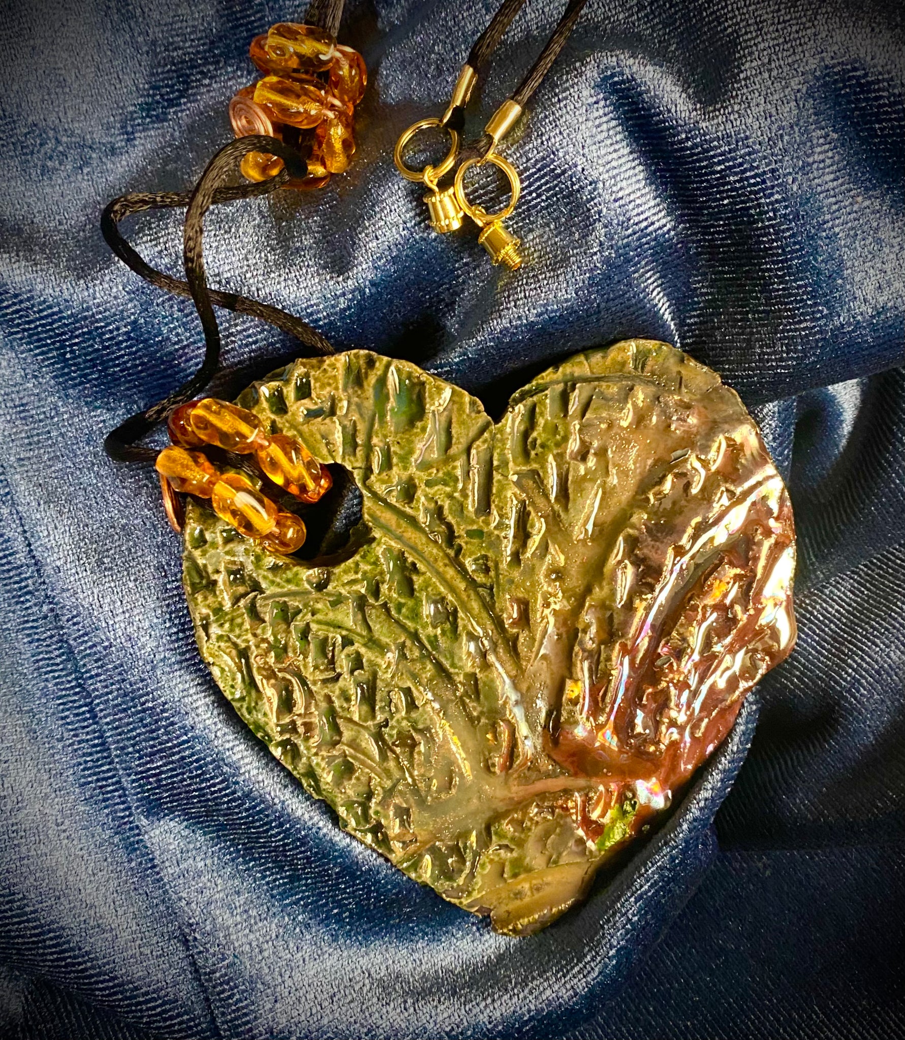 Have A Heart ! Each heart pendant is handmade with love! It is 3"x 3" and weighs approx. 3ozs. This pendant has an alligator green and gold metallic raku glazes that renders a unique translucent  patina. The heart  has a textured  pattern. It holds a spiral of amber mini beads on a spiral copper wire. This pendant has a nice 12" black suede cord!