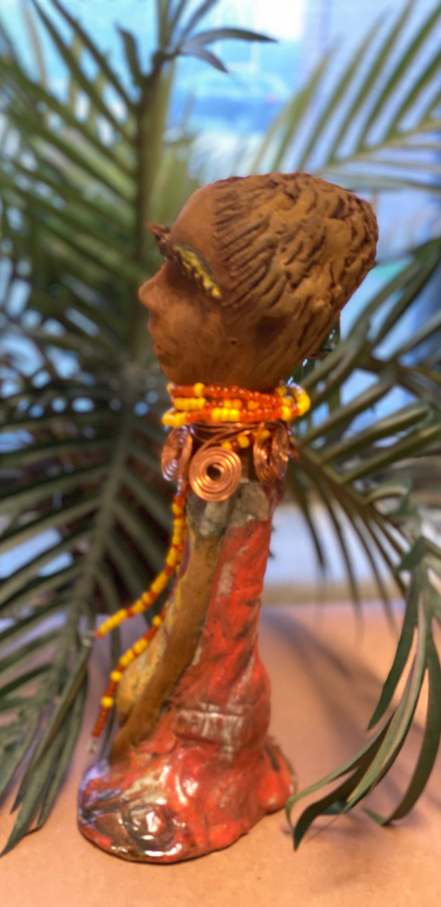 Reandra has a colorful reddish metallic green antique copper glazed dress. She wears a spiral copper wire necklace on top of a yellow red beaded collar. With eyes slightly opened, Reandra has hopes of finding a new home.