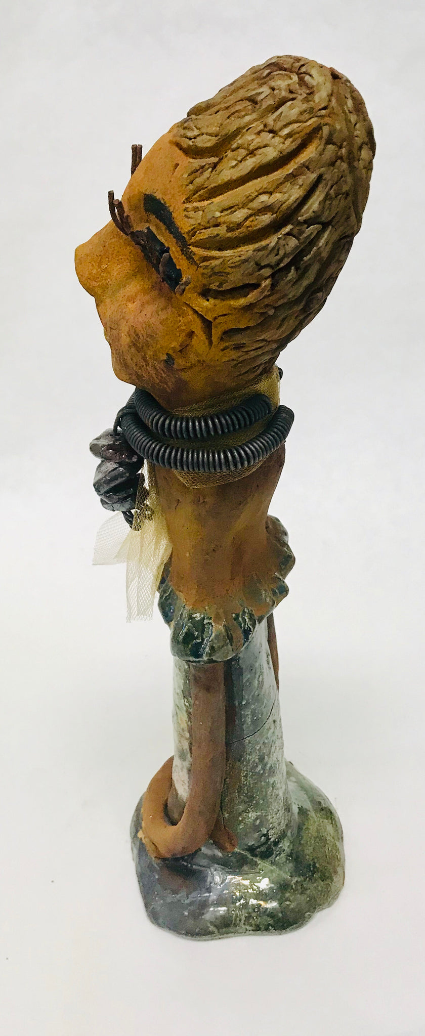 Trinity stands 14" x 4" x 4" and weighs 2.7 lbs. She has a lovely honey brown complexion with  reddish brown lips. She has a braided hairstyle.  Trinity has a colorful metallic green antique copper glazed dress. She wears a spiral wire necklaces with raku beads underneath an antique gold tulle.. With long lashes and eyes wide opened, Trinity has hopes of finding a new home.  