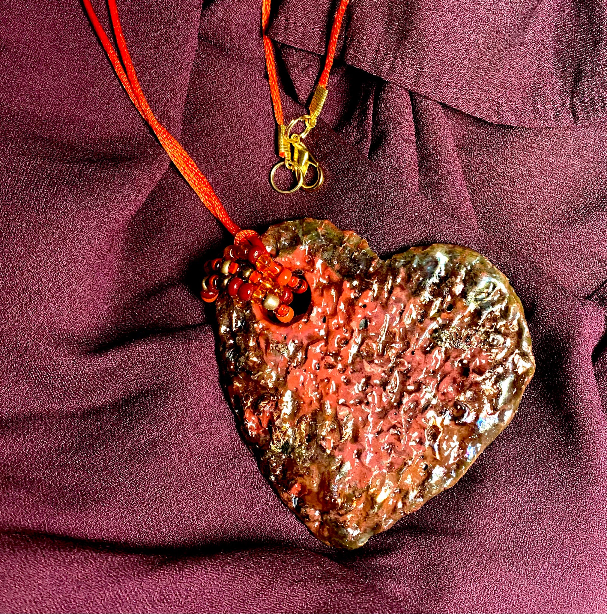 Have A Heart! Each heart pendant is handmade with love! It is 3"x 3" and weighs approx. 3ozs. This pendant has a red violet and gold metallic raku glazes that renders a unique translucent  patina. The heart has a textured pattern . Both sides are  are different and equally beautiful! It holds a spiral of red mini beads on a spiral copper wire. This pendant has a nice 12" red rattail cord!