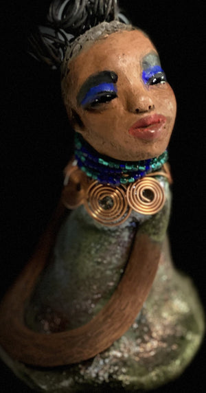Meet Nia! Nia stands 8" x 6" x 6" and weighs 1.08 lbs. She has a lovely honey brown complexion with  reddish brown lips. She has a braided  wire bun hairstyle.  Nia has a colorful metallic green antique copper glazed dress. She wears a spiral copper wire necklaces with green beads underneath.  With eyes wide opened, Nia has hopes of finding her way into your home.