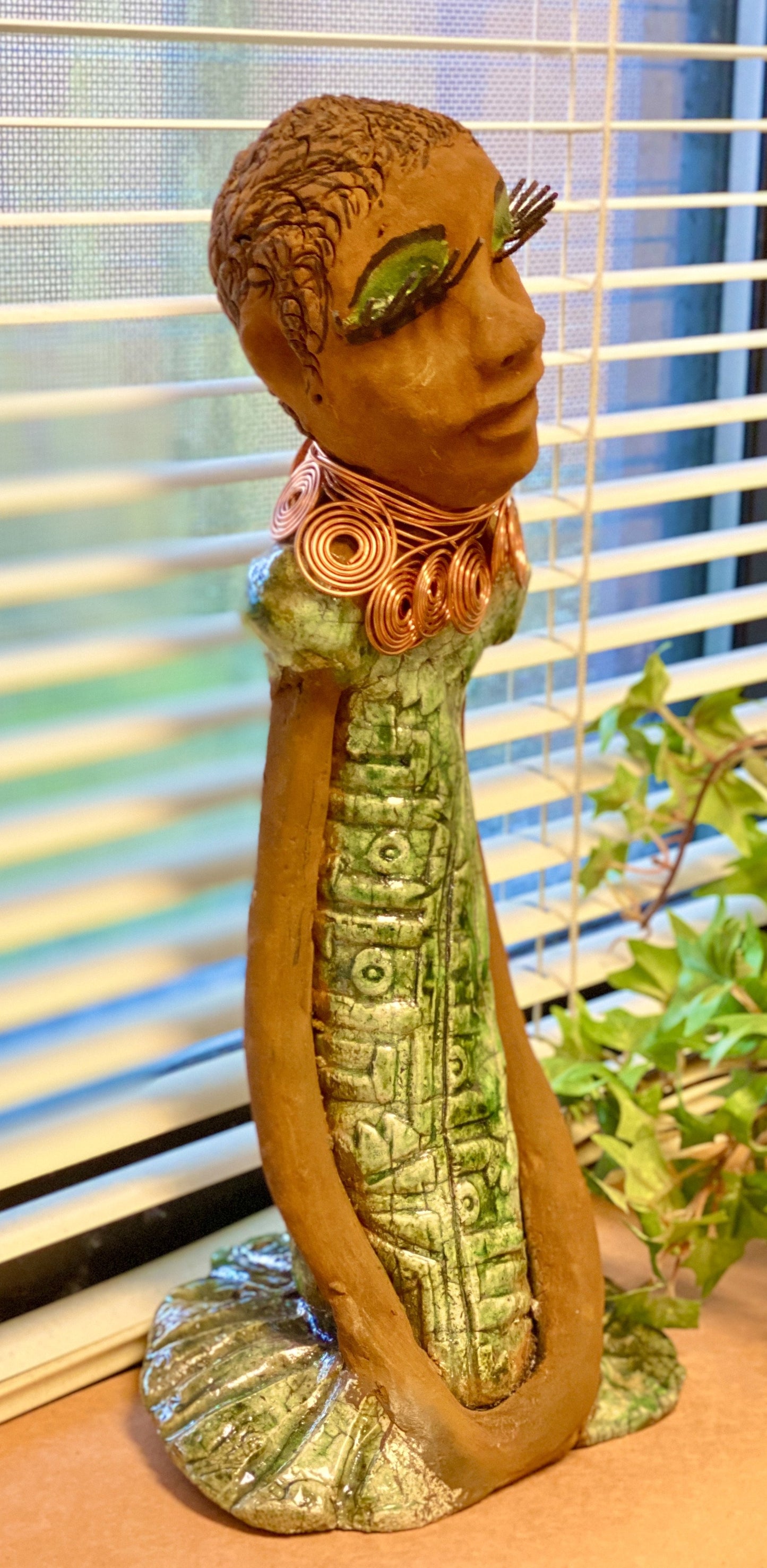 Gabrielle  stands 14" x 6" x 5.5" and weighs 2.15 lbs. She has a lovely honey brown complexion with green eye shadow. Her hairstyle is etched in clay with tribal markings.  Gabrielle  long loving arms rest beside her alligator green dress. She wears a spiral copper necklace. Gabrielle  is a sophisticated lady that will grace your home.