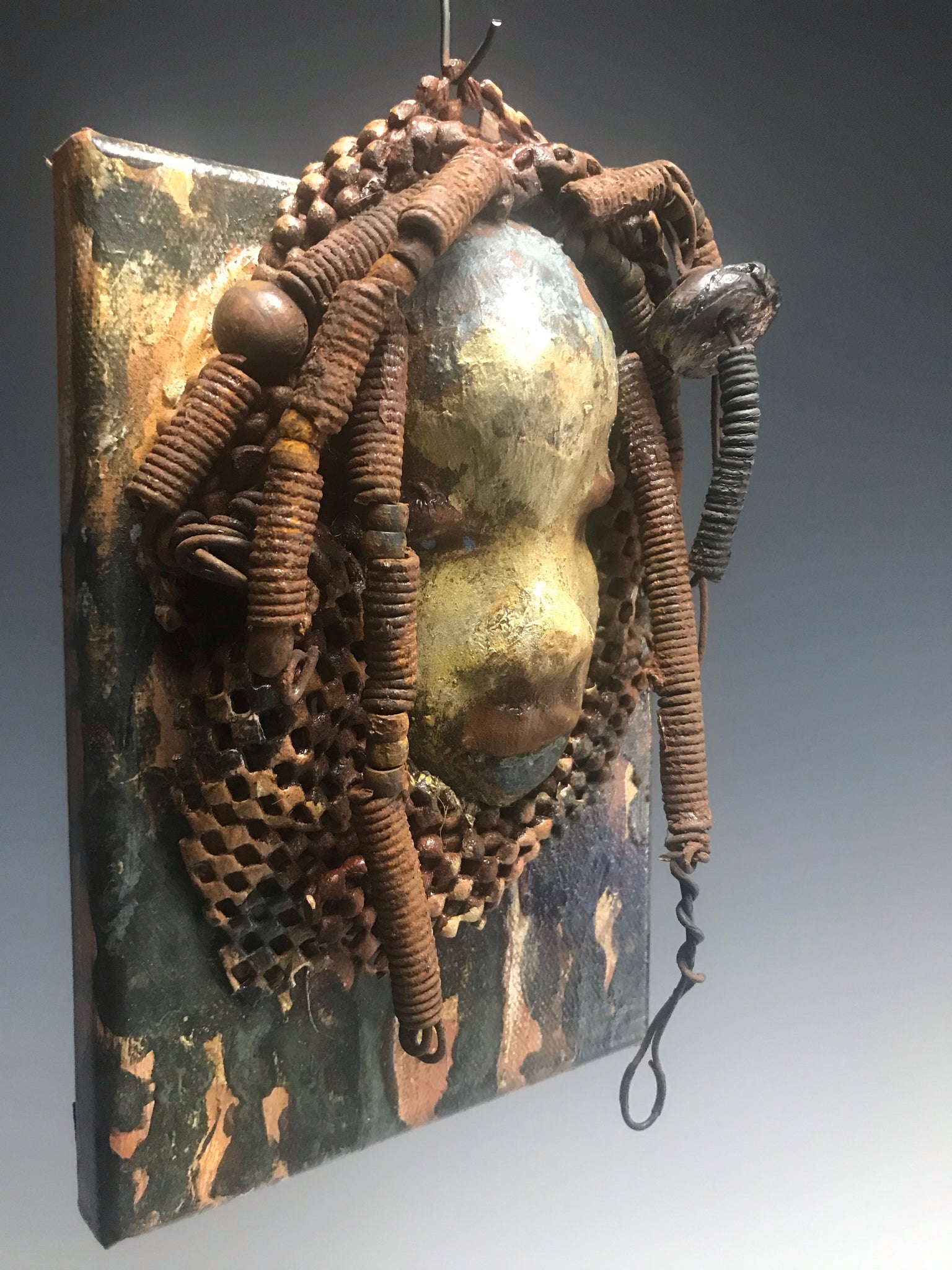 "I started making mask after seeing an authentic African Mask collection at the Smithsonian Museum of African Art. I was in total awe. Dillan was inspired by my visit there".       Dillan is mounted on a painted 6"x 4"x 2" canvas. She weighs 15 ounces.     Her face is formed with hand coiled wire, multiple raku, and wood beads, matte textured  and brown cloth.     Dillan's face beams with  almond  green glaze and rust blue brown lips.     Dillan is ready to be hung.