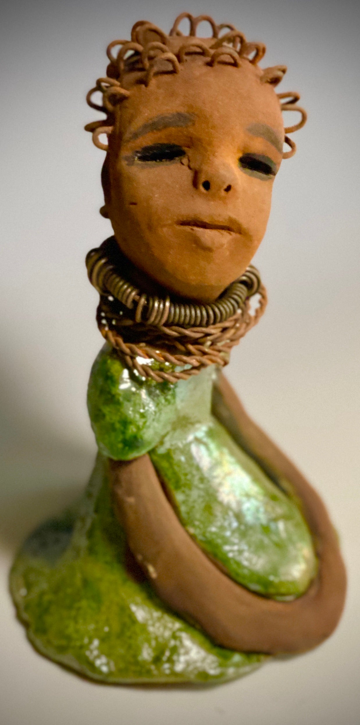 Meet Nura! Nura stands 7.5" x 5" x 5" and weighs 1.06 lbs. She has a lovely honey brown complexion with  reddish brown lips. She has a wire braided hairstyle.  Nura has a colorful metallic green antique copper glazed dress. She wears spiral wire necklaces  With her eyes wide opened, Nura has hopes of finding  her way into your  home.