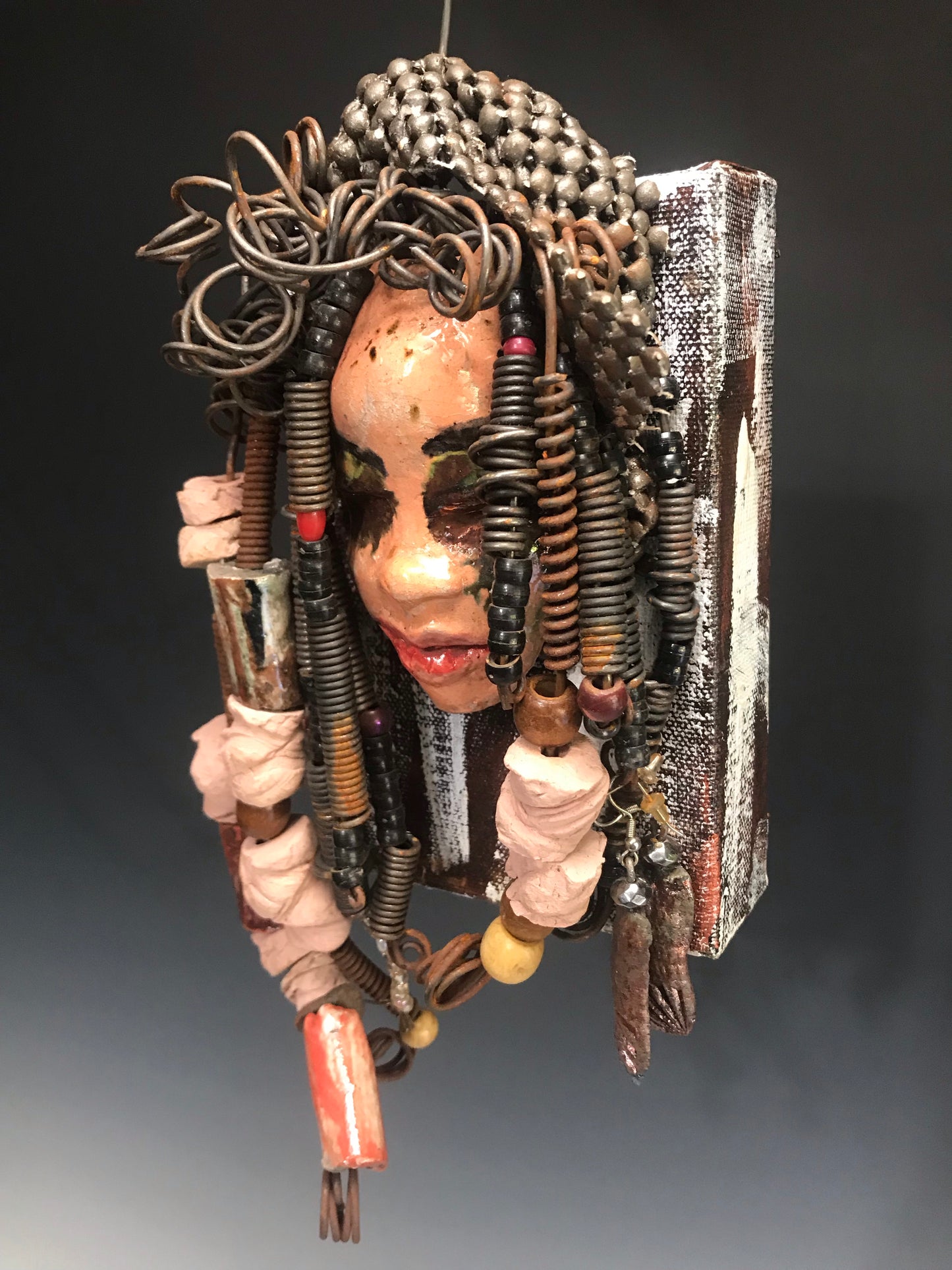      Kamara is uniquely mounted on a painted 6"x 4"x 2" canvas. It weighs 1.6lbs.     Her face is formed with hand coiled wire, raku beads, textured brown cloth, and multi color raku beads.     Kamara's face beams with pale pink crackle glaze and ruby red lips.  Kamara is ready to be hung in a special place!   