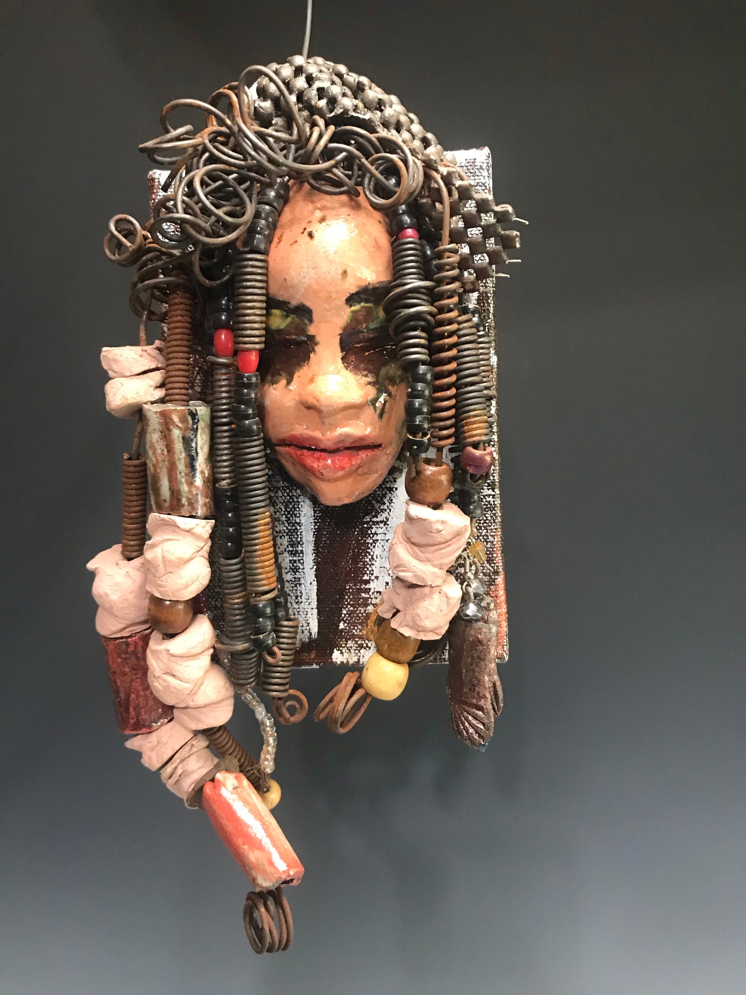      Kamara is uniquely mounted on a painted 6"x 4"x 2" canvas. It weighs 1.6lbs.     Her face is formed with hand coiled wire, raku beads, textured brown cloth, and multi color raku beads.     Kamara's face beams with pale pink crackle glaze and ruby red lips.  Kamara is ready to be hung in a special place!   