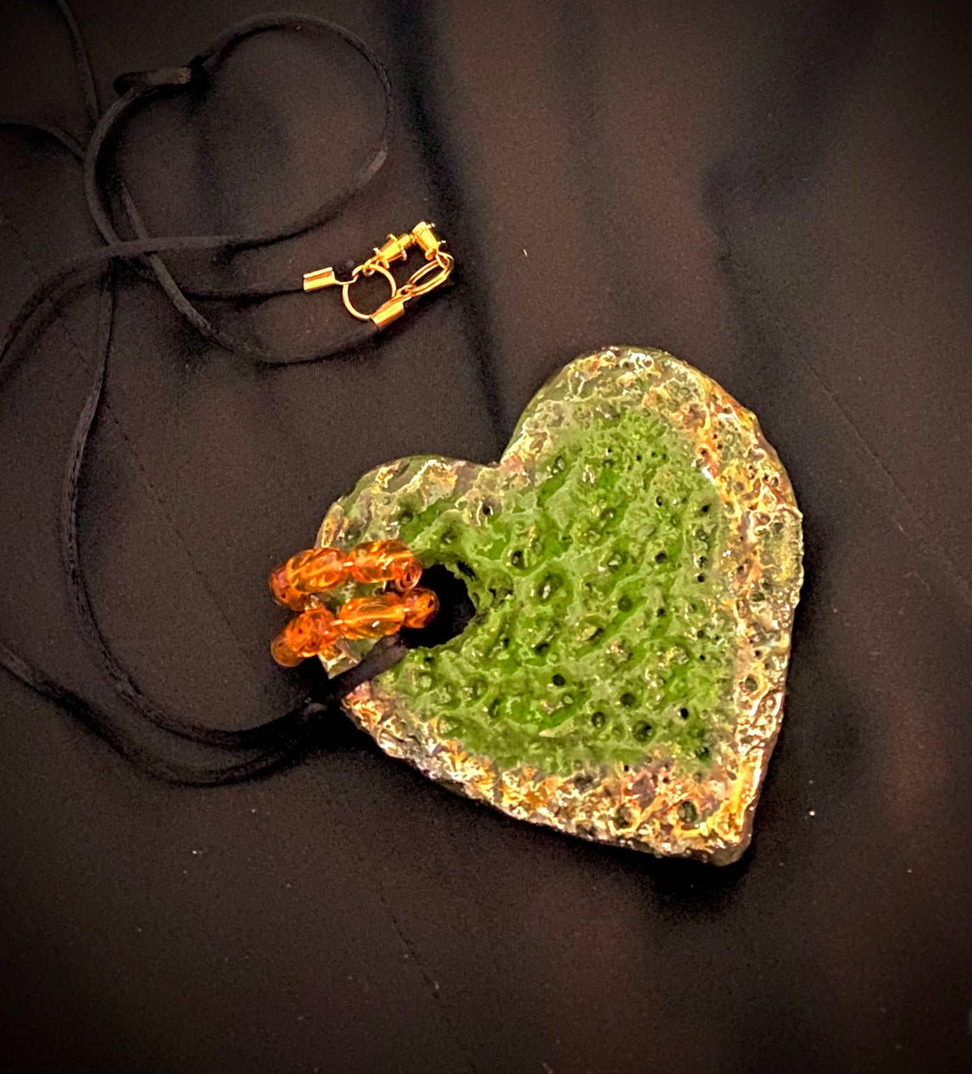 Have A Heart ! Each heart pendant is handmade with love! It is 3"x 3" and weighs approx. 3ozs. This pendant has a green and gold metallic raku glazes that renders a unique translucent  patina. The heart is highly textured. It holds a spiral of amber mini beads on a spiral copper wire. This pendant has a nice 12" black suede cord!