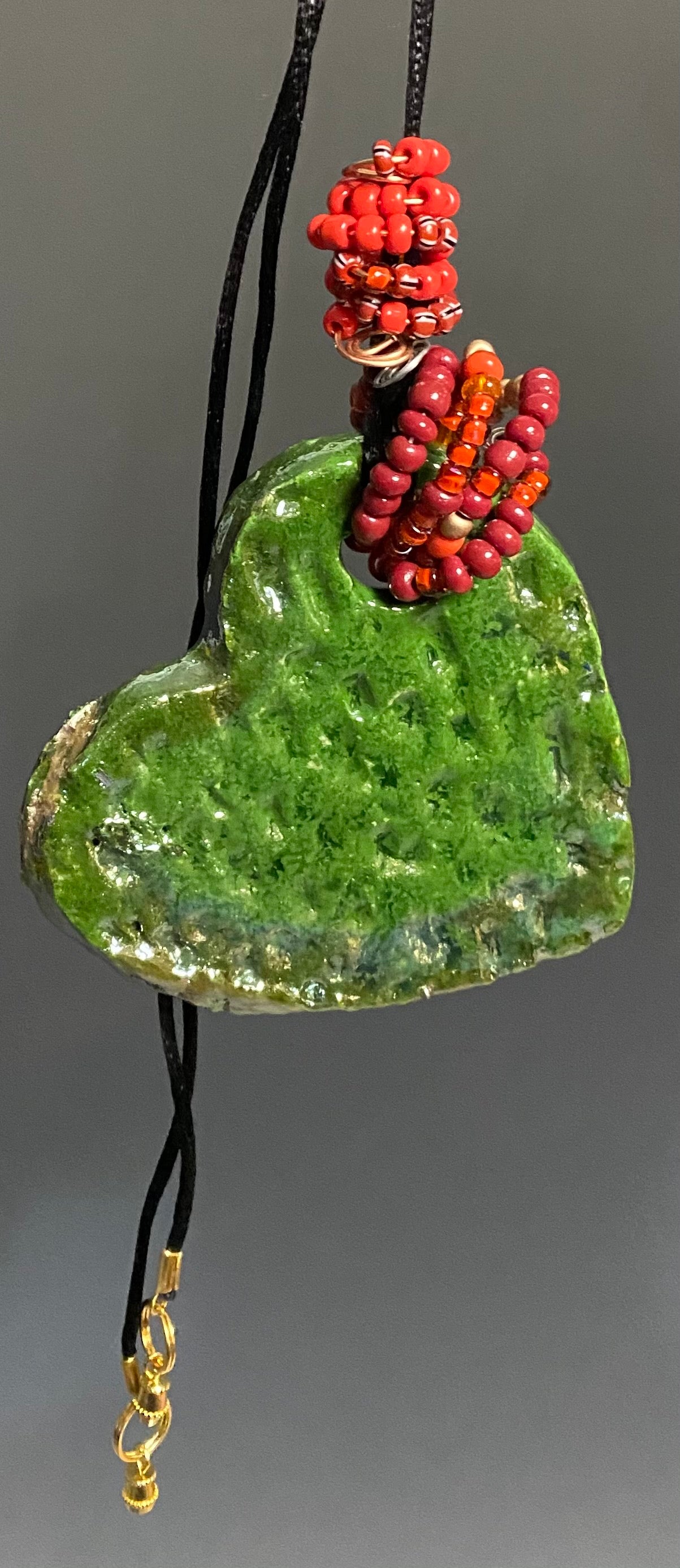  Have A Heart! Each heart pendant is handmade with love! It is 3"x 3"and weighs approx. 3ozs. This pendant has a emerald green and gold metallic raku glazes that renders a unique translucent  patina. The heart has a textured pattern . Both sides are  are different and equally beautiful! It holds a spiral of red  and orange mini beads on a spiral copper wire. This pendant has a nice 12" black adjustable rattail cord!