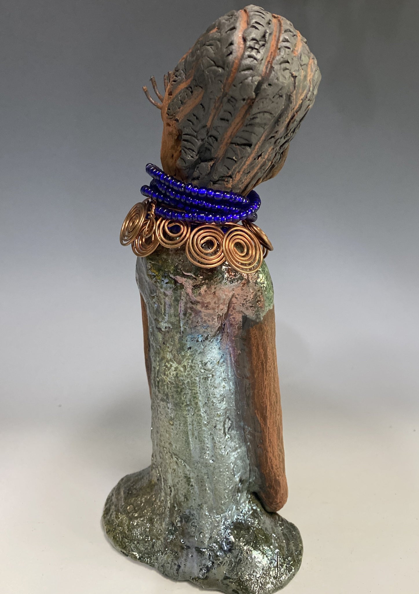 Meet Ophelia! Ophelia stands 7.5" x 4" x 5" and weighs 1.02  lbs. She has a lovely honey brown complexion with reddish brown lips. She has a short braided hairstyle.  Ophelia has a colorful metallic antique copper glazed dress. She wears spiral copper wire necklaces on top of an aqua blue beaded collar. Ophelia long loving arms rest at her side. With accent blue eye shadow and eyes wide opened, Ophelia has hopes of finding  her way into your home!