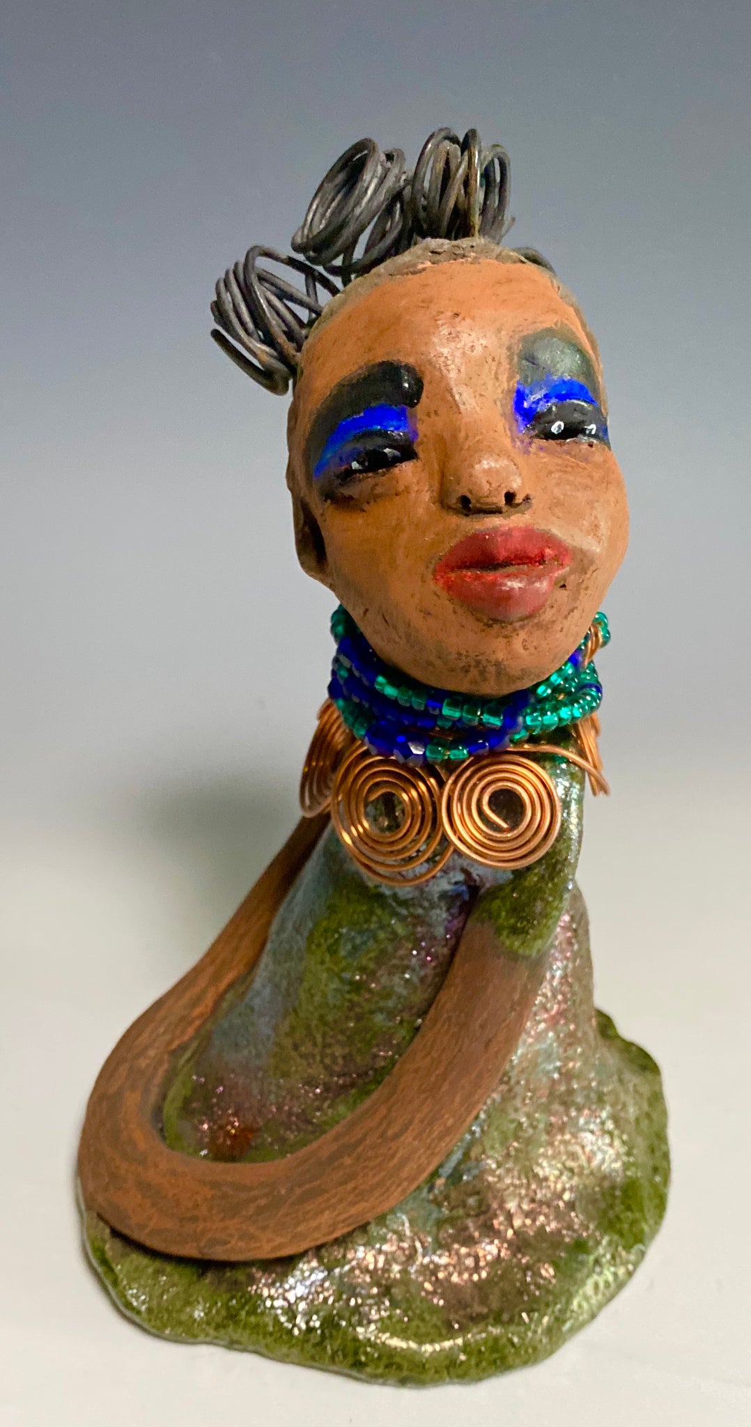 Meet Nia! Nia stands 8" x 6" x 6" and weighs 1.08 lbs. She has a lovely honey brown complexion with  reddish brown lips. She has a braided  wire bun hairstyle.  Nia has a colorful metallic green antique copper glazed dress. She wears a spiral copper wire necklaces with green beads underneath.  With eyes wide opened, Nia has hopes of finding her way into your home.