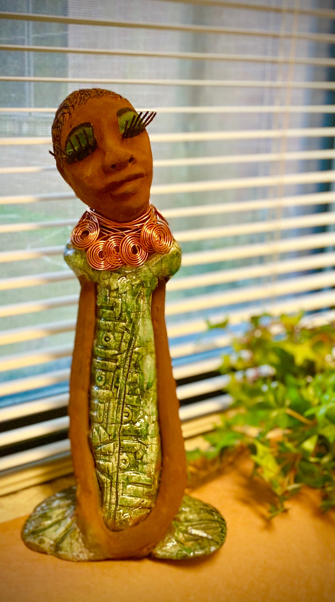 Gabrielle  stands 14" x 6" x 5.5" and weighs 2.15 lbs. She has a lovely honey brown complexion with green eye shadow. Her hairstyle is etched in clay with tribal markings.  Gabrielle  long loving arms rest beside her alligator green dress. She wears a spiral copper necklace. Gabrielle  is a sophisticated lady that will grace your home.