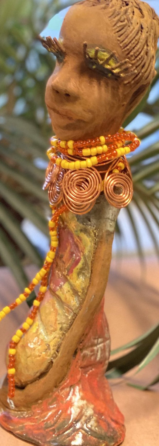 Reandra has a colorful reddish metallic green antique copper glazed dress. She wears a spiral copper wire necklace on top of a yellow red beaded collar. With eyes slightly opened, Reandra has hopes of finding a new home.