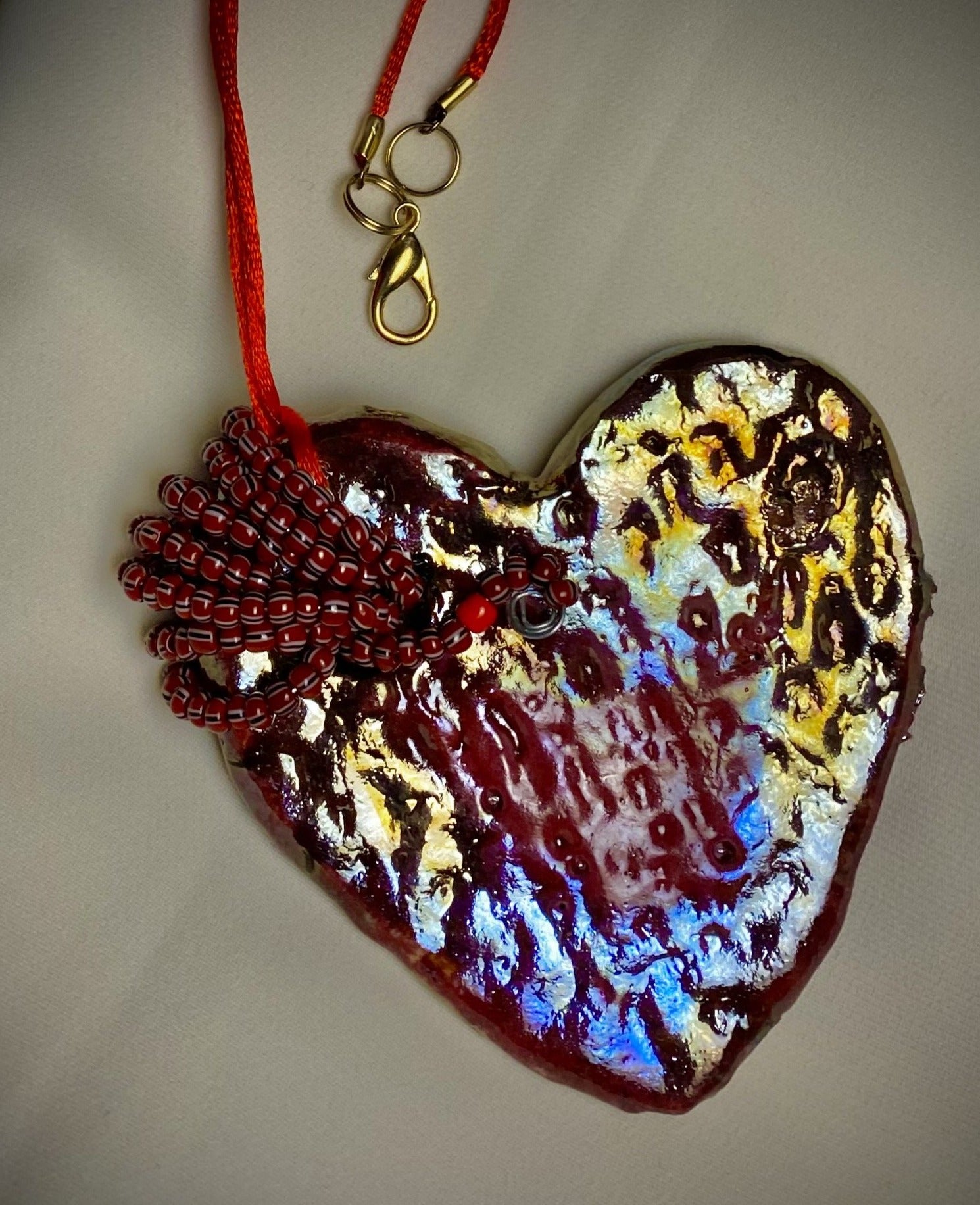 Have A Heart ! Each heart pendant is handmade with love! It is 3"x 3" and weighs approx. 3ozs. This pendant has a red violet and gold metallic raku glazes that renders a unique translucent  patina. The heart has a textured pattern . Both sides are  are different and equally beautiful! It holds a spiral of red violet mini beads on a spiral copper wire. This pendant has a nice 12" red suede cord!