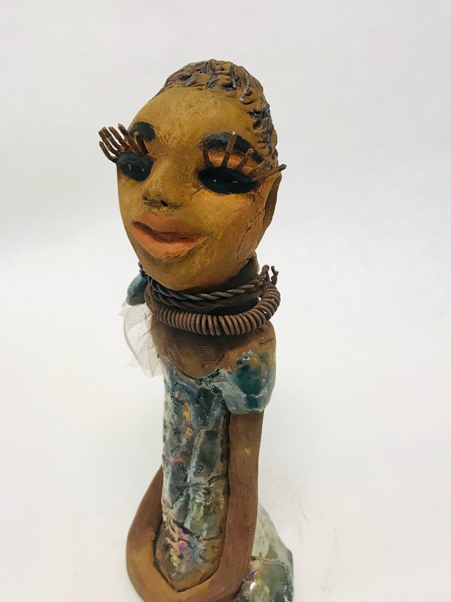 Sorbari stands 11" x 4" x 4" and weighs 2.5 lbs. She has a lovely honey brown complexion with  reddish brown lips. She has a braided hairstyle.  Sorbari has a colorful metallic green antique copper glazed dress. She wears two spiral wire necklaces. With long eyes slightly opened, Sorbari has hopes of finding a new home.  