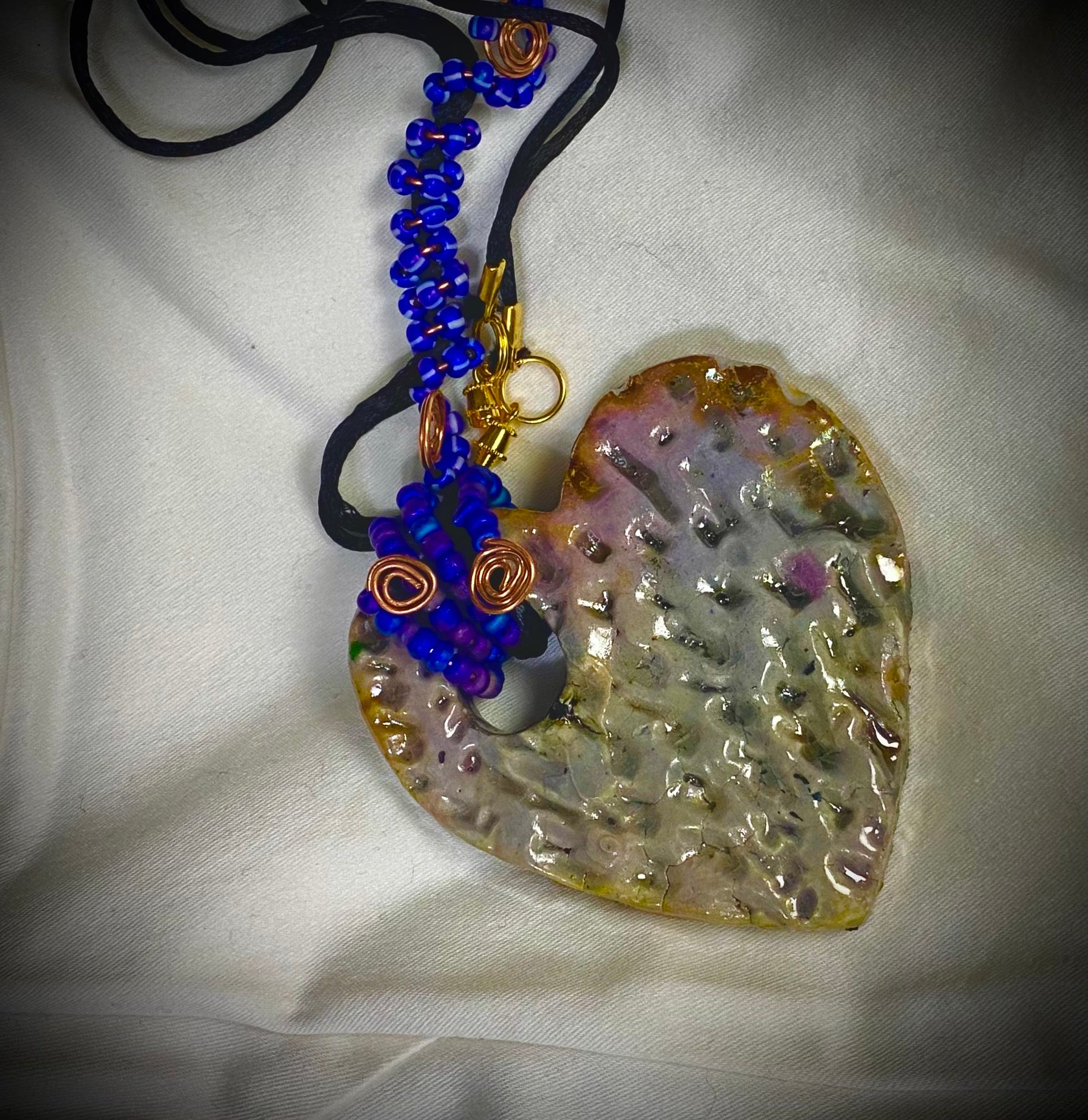   Have A Heart ! Each heart pendant is handmade with love! It is 3"x 3" and weighs approx. 3ozs. This pendant has a white metallic raku glazes that renders a unique translucent  patina. The heart has a textured pattern . It holds a spiral of blue mini beads on a spiral copper wire. This pendant has a nice 12" black suede cord!