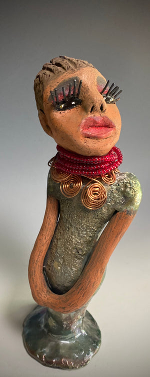 Meet Mona! Mona stands 11.5" x 4" x 4" and weighs 1.81 lbs. She has a lovely chocolate brown complexion with chocolate brown lips. She has a braided hairstyle.  Mona has a colorful metallic antique copper glazed dress. She wears a spiral copper wire necklaces on top of a beaded ruby red collar. With long lashes and eyes wide opened, Mona has hopes of finding a new home. You may have noticed, Mona is the one of a few herdew  sculptures that wears a face  covering during this unprecedented time.
