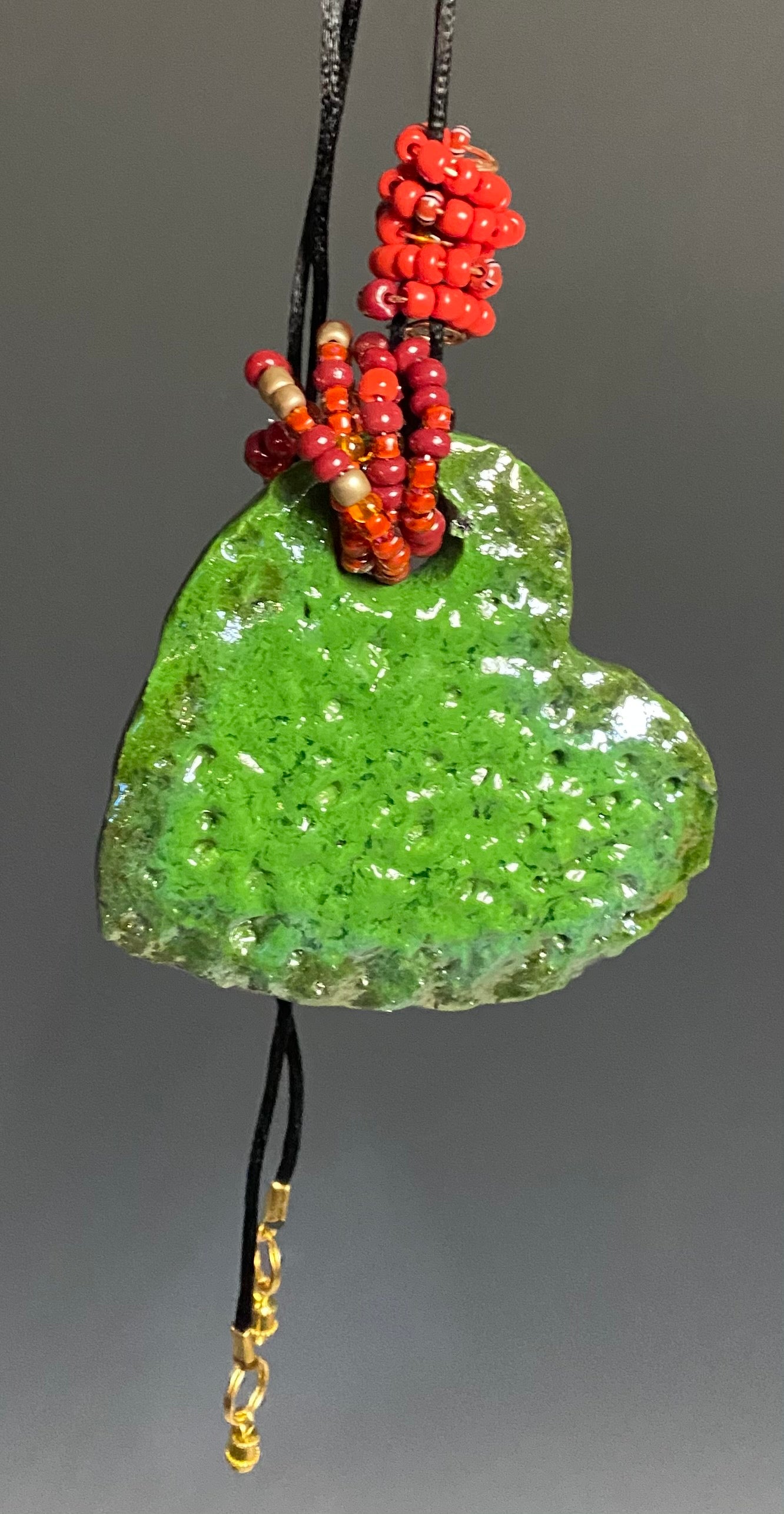  Have A Heart! Each heart pendant is handmade with love! It is 3"x 3"and weighs approx. 3ozs. This pendant has a emerald green and gold metallic raku glazes that renders a unique translucent  patina. The heart has a textured pattern . Both sides are  are different and equally beautiful! It holds a spiral of red  and orange mini beads on a spiral copper wire. This pendant has a nice 12" black adjustable rattail cord!