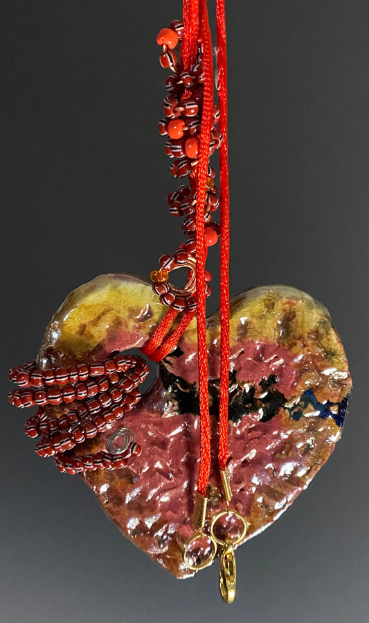 Have A Heart! Each heart pendant is handmade with love! It is 3"x 3"and weighs approx. 3ozs. This pendant has a red, white, and gold metallic raku glazes that renders a unique translucent  patina. The heart has a textured pattern . Both sides are  are different and equally beautiful! It holds a spiral of red  and deep purple mini beads on a spiral copper wire. This pendant has a nice 12" red adjustable rattail cord!