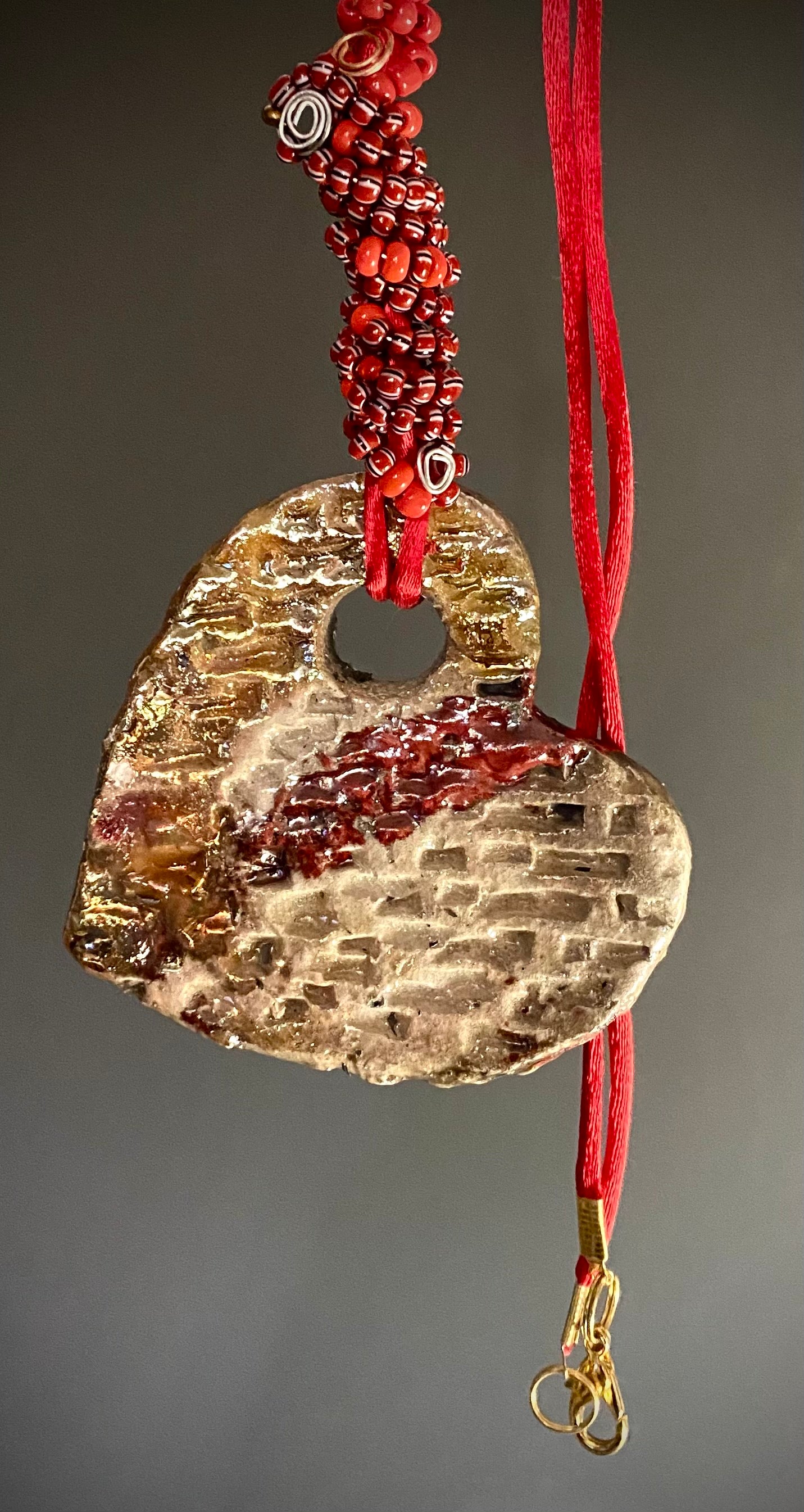Have A Heart! Each heart pendant is handmade with love! It is 3"x 3"and weighs approx. 3ozs. This pendant has a red, white, and gold metallic raku glazes that renders a unique translucent  patina. The heart has a textured pattern . Both sides are  are different and equally beautiful! It holds a spiral of red and deep purple mini beads on a spiral copper wire. This pendant has a nice 12" red adjustable rattail cord!
