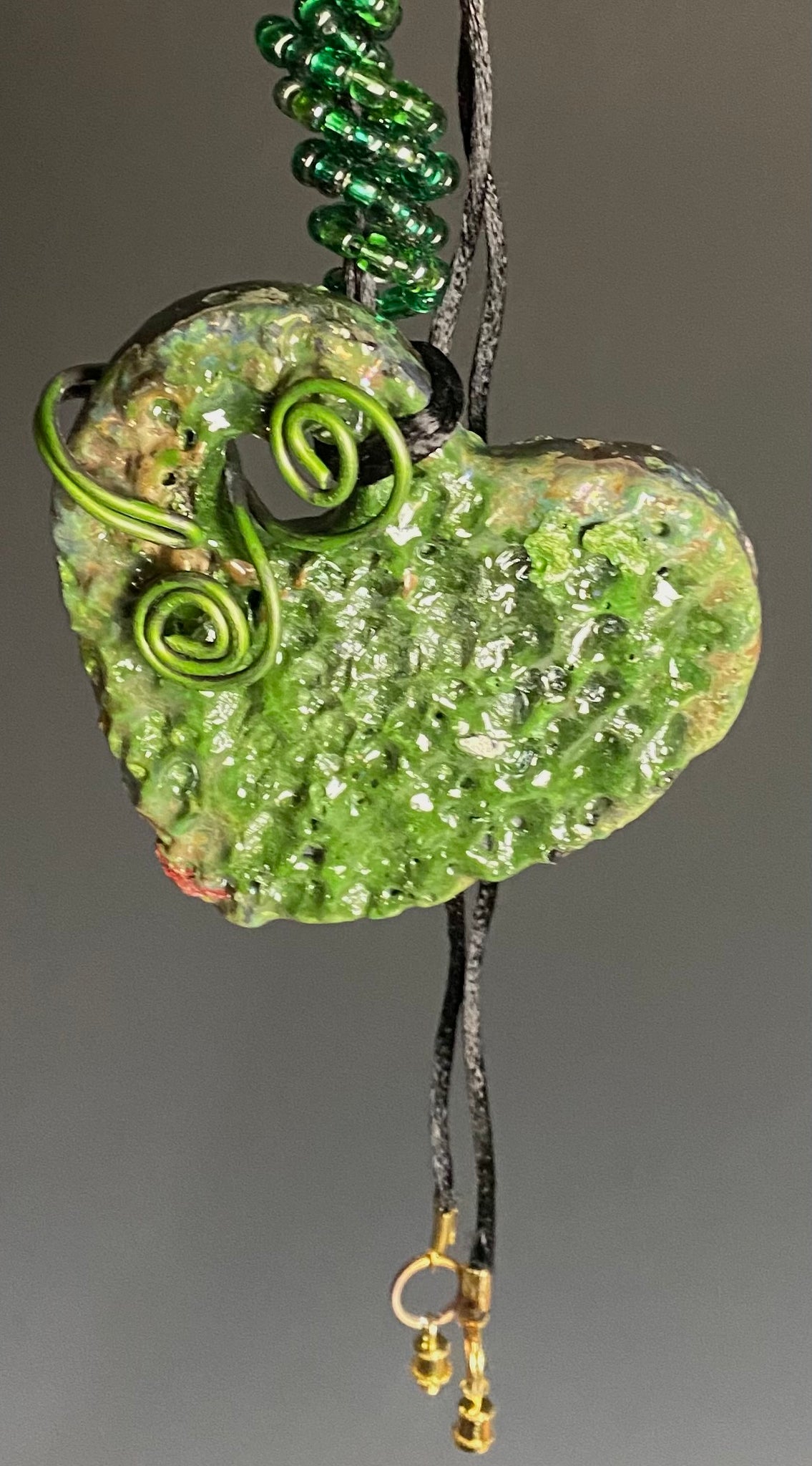 Have A Heart! Each heart pendant is handmade with love! It is 3"x 3"and weighs approx. 3ozs. This pendant has a green and  gold metallic raku glazes that renders a unique translucent  patina. The heart has a textured pattern . Both sides are  are different and equally beautiful! It holds a spiral of green mini beads on a spiral copper wire. This pendant has a nice 12" black rattail cord!