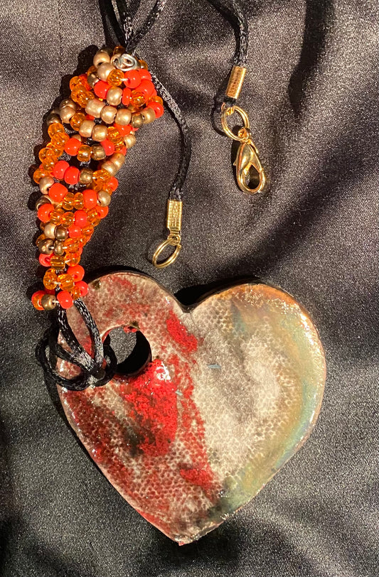 Have A Heart! Each heart pendant is handmade with love! It is 3"x 3"and weighs approx. 3ozs. This pendant has a red violet and gold metallic raku glazes that renders a unique translucent  patina. The heart has a textured pattern . Both sides are  are different and equally beautiful! It holds a spiral of red ad amber mini beads on a spiral copper wire. This pendant has a nice 12" black rattail cord!