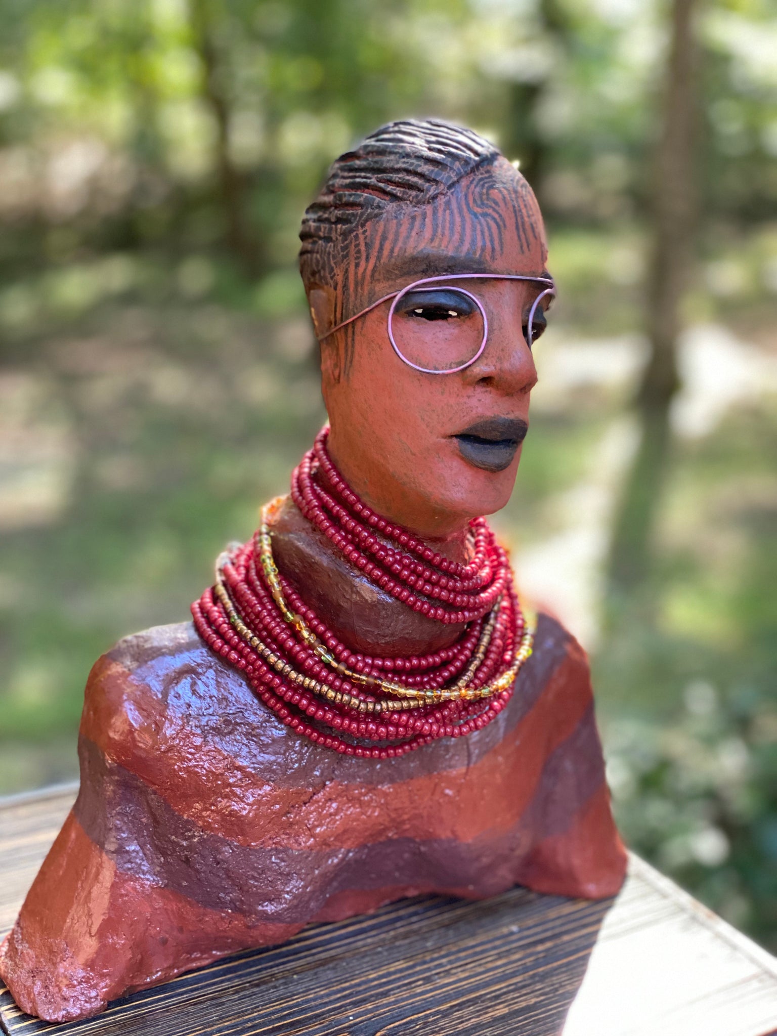 Meet Harriet! Harriet stands 13" x 11" x 5" and weighs 5 lbs. She wears a coral striped dress with a string of ruby red and gold beads. Harriet has a honey brown rust complexion  and distinct eyes. Her eyes  are slightly opened underneath her glasses... She has mixed black and copper streaks of straight corn rolls of clay hair. She has a straight look of concern as to what is about to take place.
